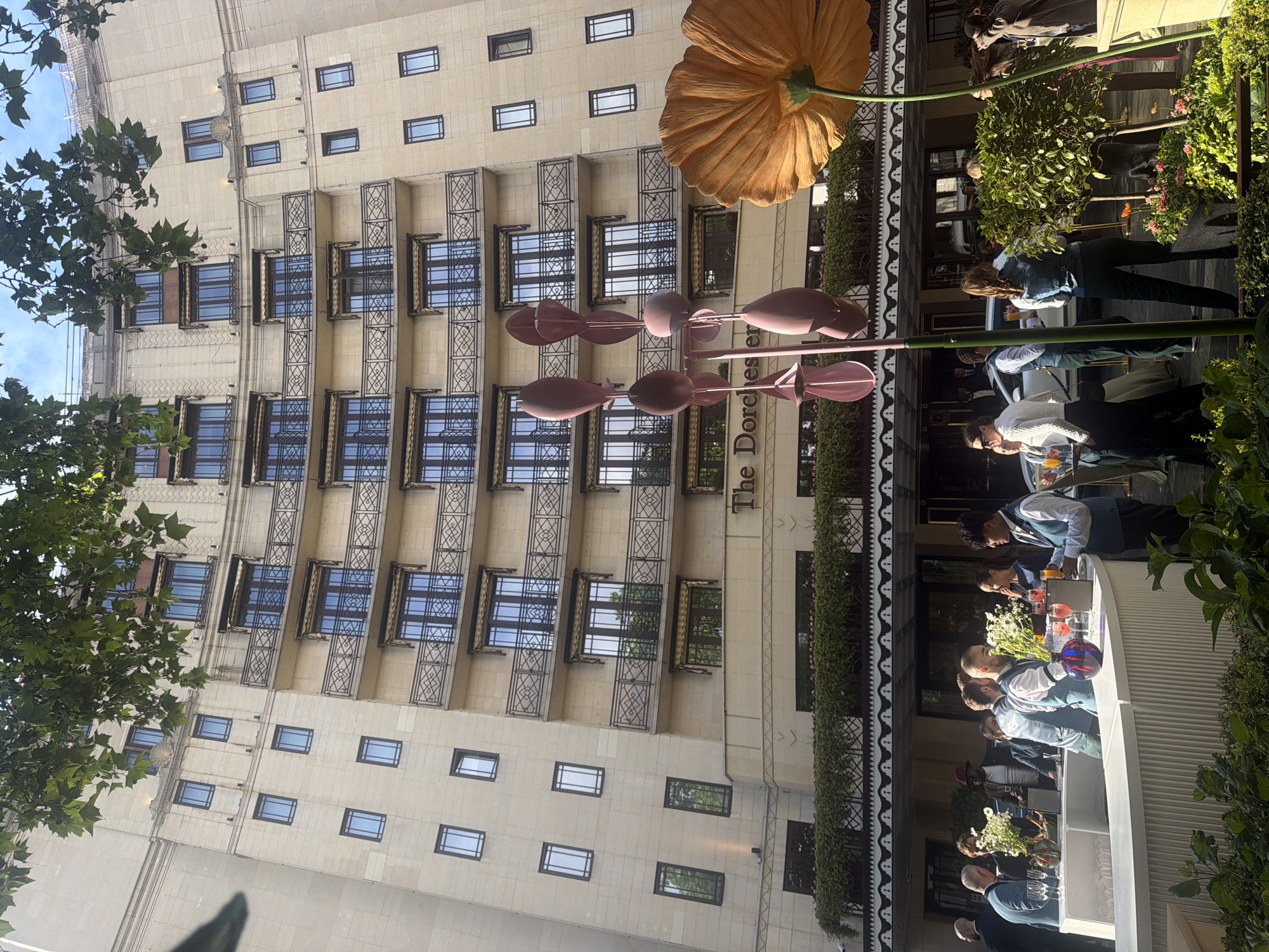 The image captures the exterior of The Dorchester, a luxury hotel in London, United Kingdom. The building has a classical façade with multiple floors, each featuring symmetrically placed windows and decorative ironwork balconies. A large artificial orange flower and a set of decorative pink wind spinners adorn the entrance area.
To the right of the frame, an outdoor bar is set up. Several staff members are visible behind the bar, preparing and serving drinks to unseen customers. The bar is decorated with white flowers, and glassware filled with colorful drinks is prominent. Lush greenery surrounds the outdoor bar area, and people are seen in the background. The image conveys a scene of upscale hospitality and urban elegance in central London.
