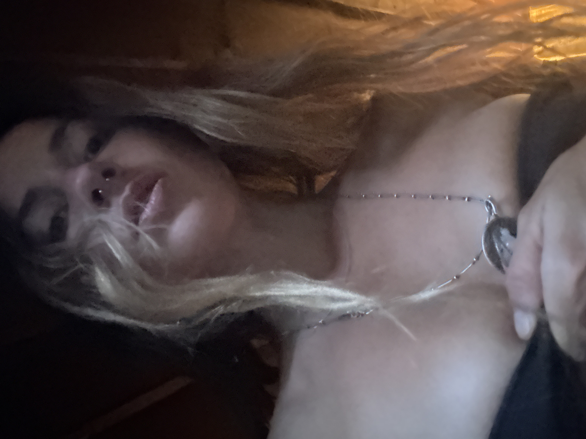 The image is a low-angle, close-up, indoor shot of a woman from the chest up. She has long, wavy blonde hair that is partially obscuring her face. Her head is tilted back, and she is looking down and slightly to her left. She is wearing a dark-colored garment that is partially visible, and a silver necklace with a round pendant. There is a nose ring visible on her right nostril. The lighting is dim, with warm tones and shadows. The background is dark and indistinct, suggesting an indoor setting. No city or specific location is discernible.