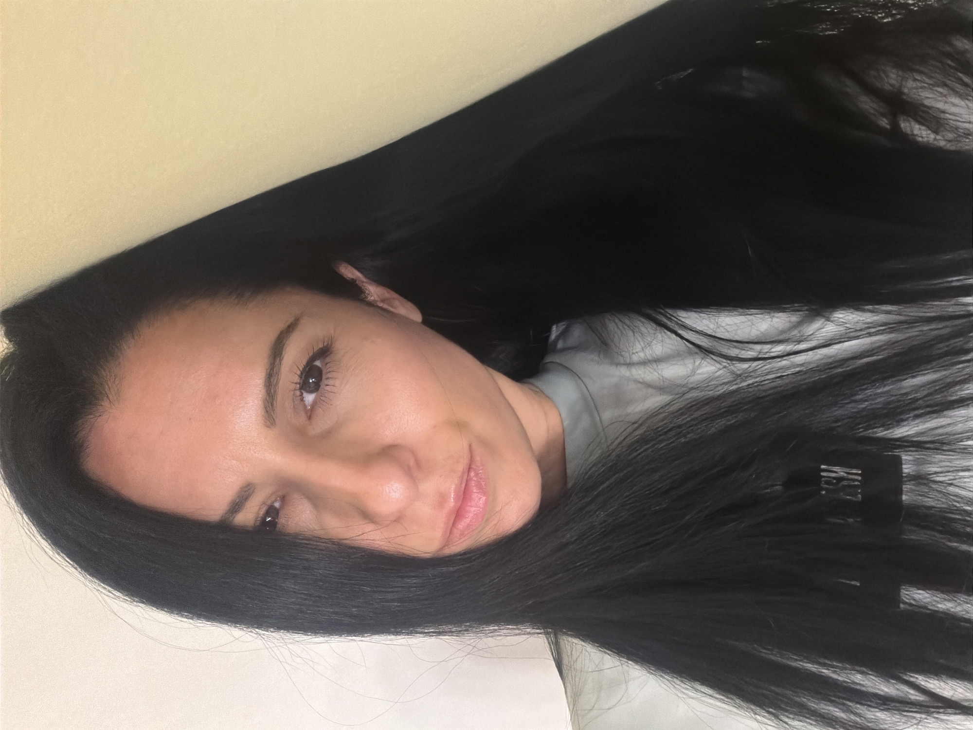 A woman with long black hair is posing for a picture. She is wearing a grey shirt. The background consists of a neutral-colored wall. The image is rotated. The location is not discernible from the image.