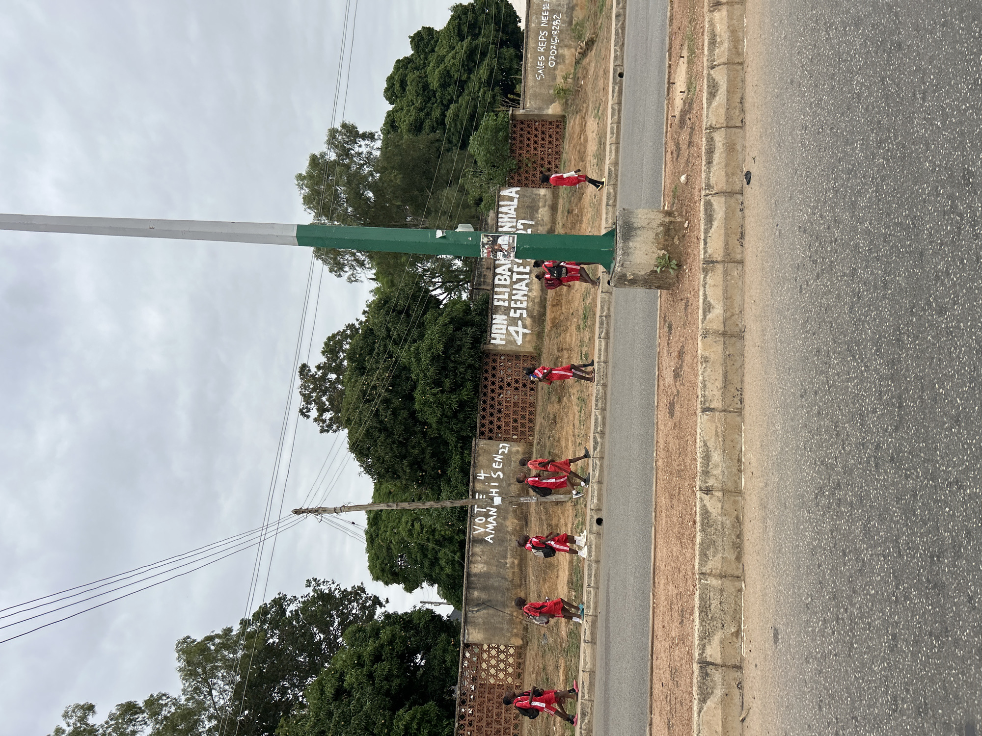 Along a roadside in Jos, Nigeria, a group of uniformed children walk past a low wall covered with political campaign graffiti. The sky is overcast. Trees are visible beyond the wall. A utility pole stands tall along the wall.