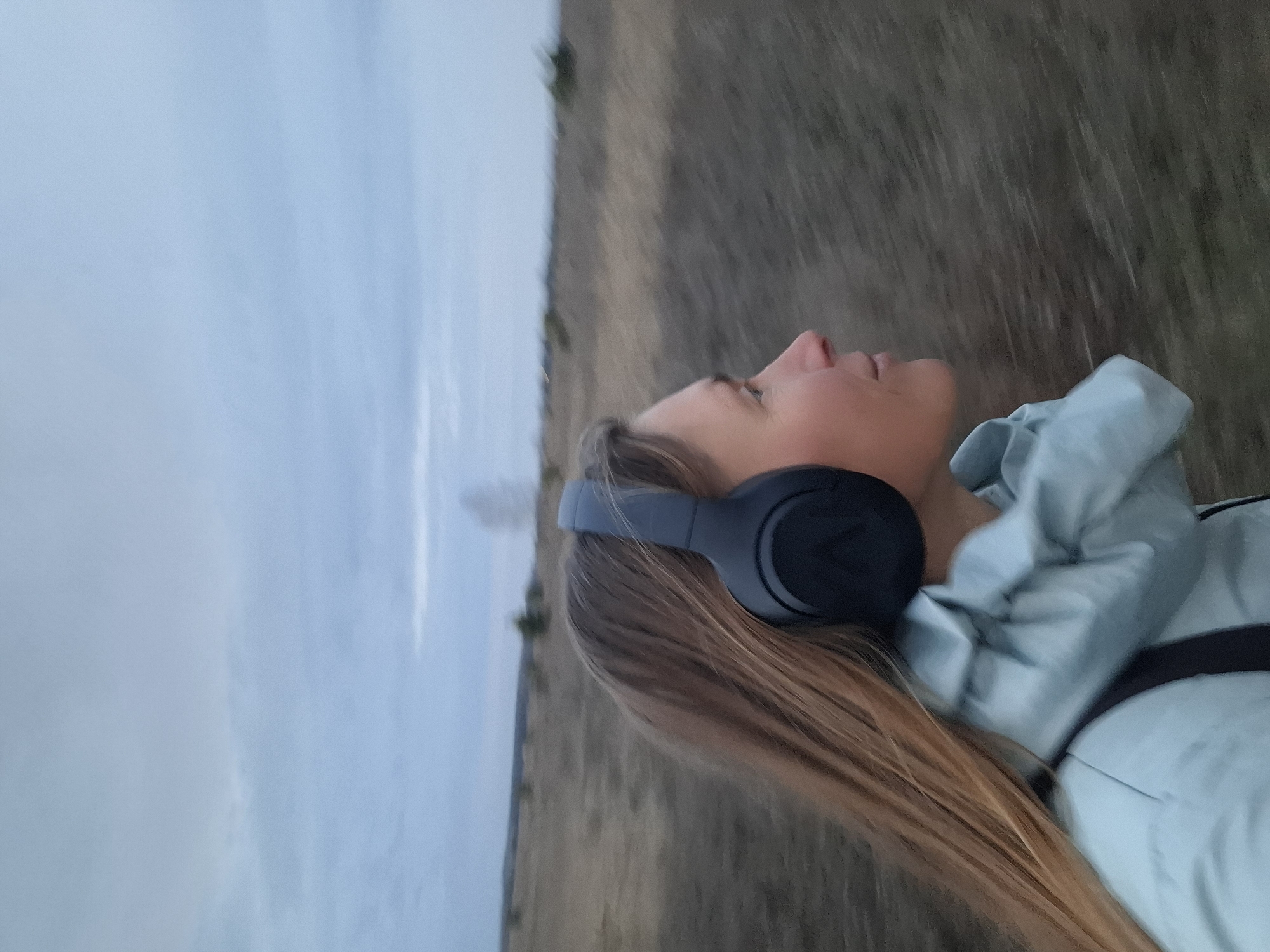 A woman with long, light brown hair is wearing headphones and a light blue jacket. She is looking up. The background consists of a field of tall, dry grass and a cloudy sky. The location is outdoors, possibly in or near the city of Khlevnoe, Russia, although this cannot be definitively determined from the image itself. There is no visible interaction in the image. The focus is on the individual and the natural environment surrounding her.