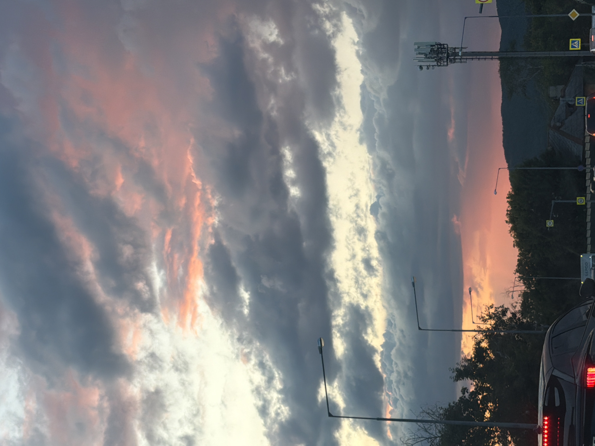 The image shows a cloudy sky with streaks of pink and orange, suggesting either sunrise or sunset. Several streetlights are visible, along with a cell tower. Trees line the horizon, and a dark-colored car is in the foreground, with its taillights illuminated. The scene appears to be a roadside view, possibly from a vehicle, with traffic signals and road signs in the background. The overall impression is of a somewhat overcast sky during twilight, with urban infrastructure present. Without specific landmarks or context, it is impossible to determine the exact location within Samara, Russia.
