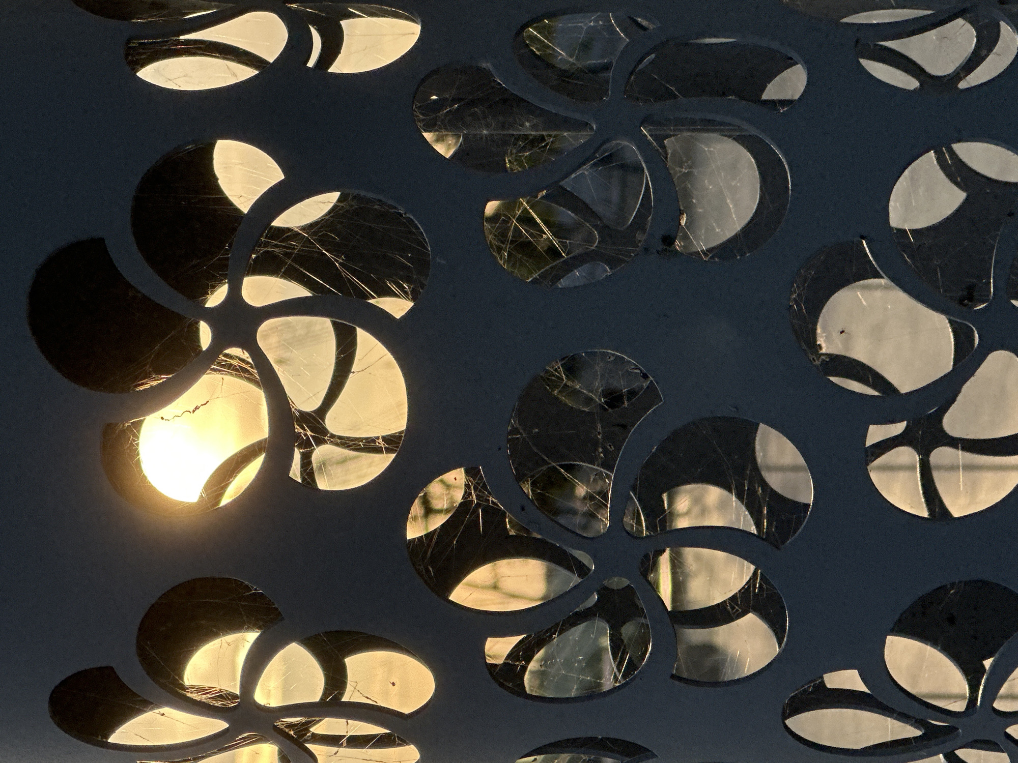 The image is a close-up shot of a dark blue, perforated metal structure, possibly a lamppost or a decorative element. The perforations are in a floral or circular pattern, and through them, the warm, golden light of the sun is visible, creating a backlit effect. The sunlight appears to be setting or rising, casting a hazy glow. Fine, cobweb-like strands are visible within the perforations, catching the light and adding a textured detail. The background visible through the holes is indistinct, showing faint shapes that suggest trees or foliage, also illuminated by the low sun. The overall impression is one of an abstract design element set against a natural, luminous backdrop. Without further context or identifying markers, the specific location within Saint Petersburg, Russia, cannot be determined from the image alone.