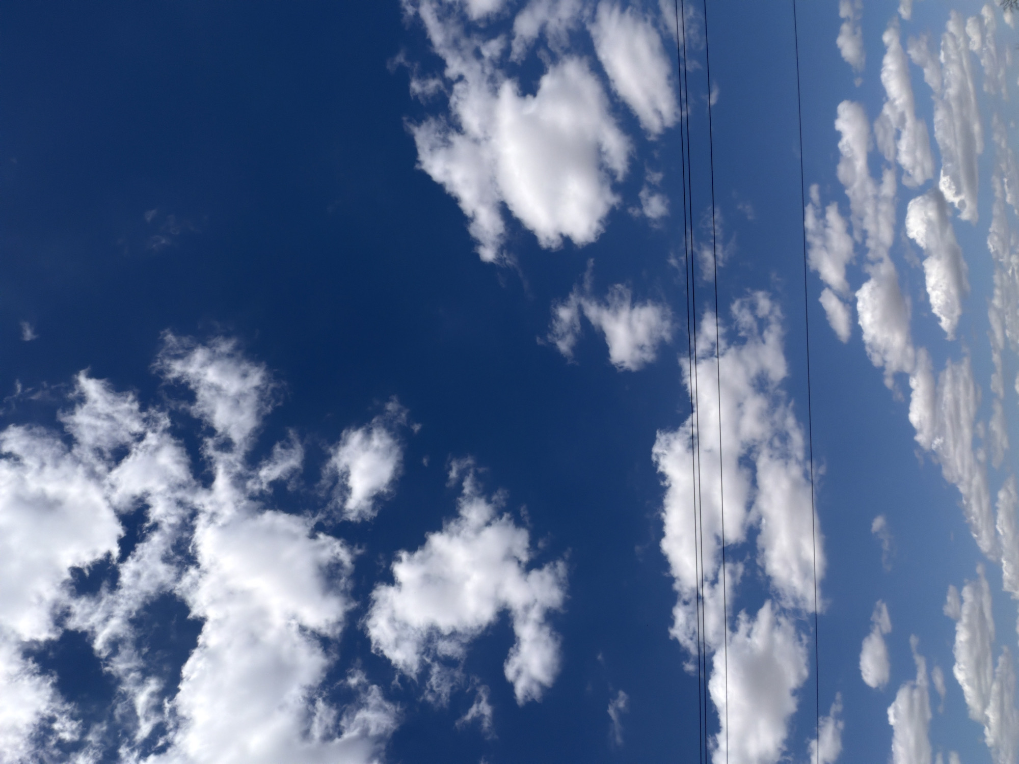 The image shows a bright blue sky filled with scattered white cumulus clouds. In the lower portion of the frame, three dark, thin horizontal lines stretch across the scene, indicating power or utility lines. There are no people, buildings, or any other discernible objects or environmental features visible in this image. The sky appears clear and sunny, suggesting fair weather conditions. This scene could be observed in any open area with a clear view of the sky, including within the city of Krasnyi Krym, Russia, particularly in areas with fewer tall structures or trees that might obstruct the view.