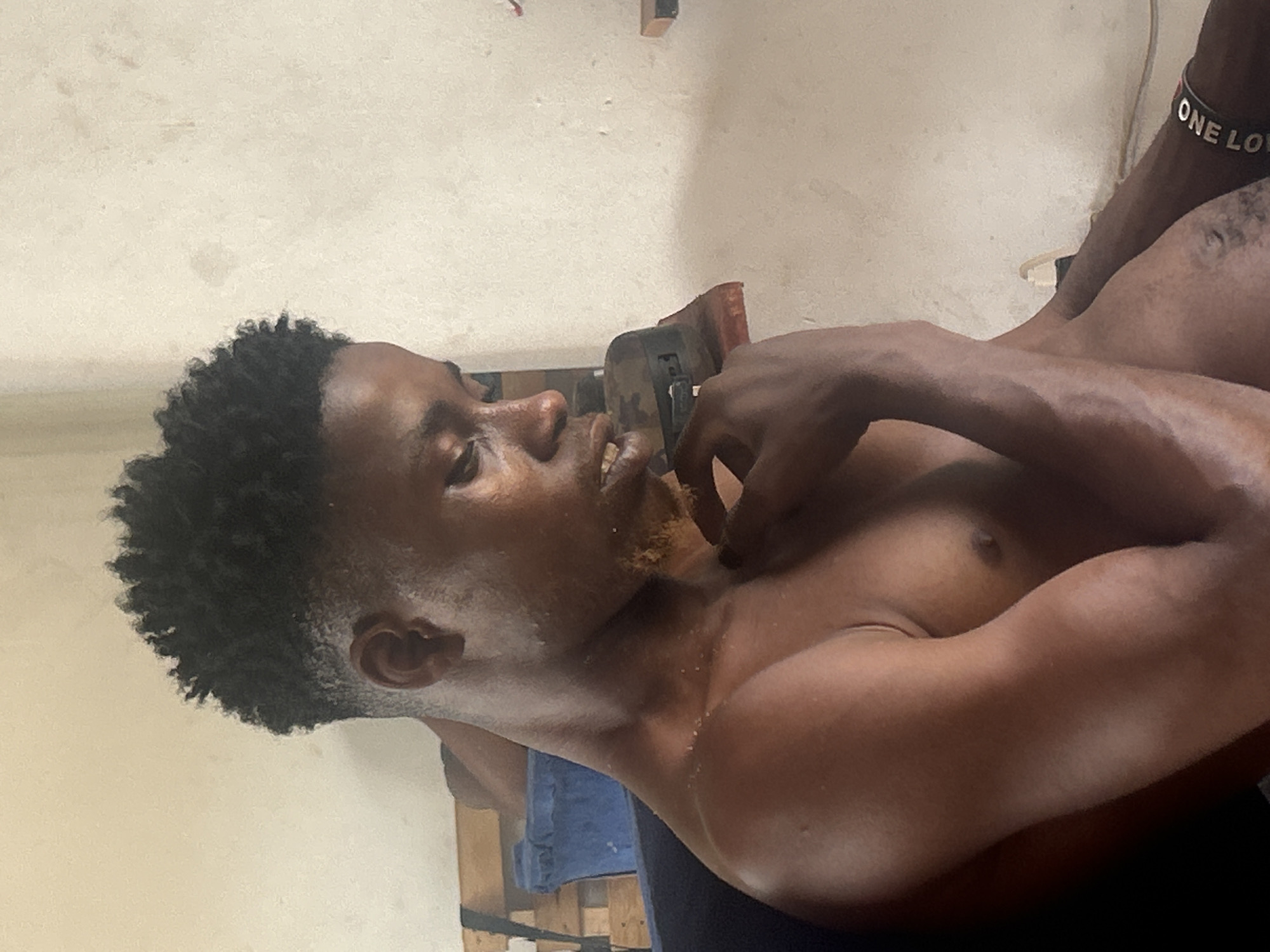 The image shows a shirtless man with dark curly hair lying on his back. His head is turned to the left, and his right hand is near his neck. He is wearing a bracelet that reads "ONE LOVE" on his right wrist. The background appears to be a plain wall. The location is unknown.
