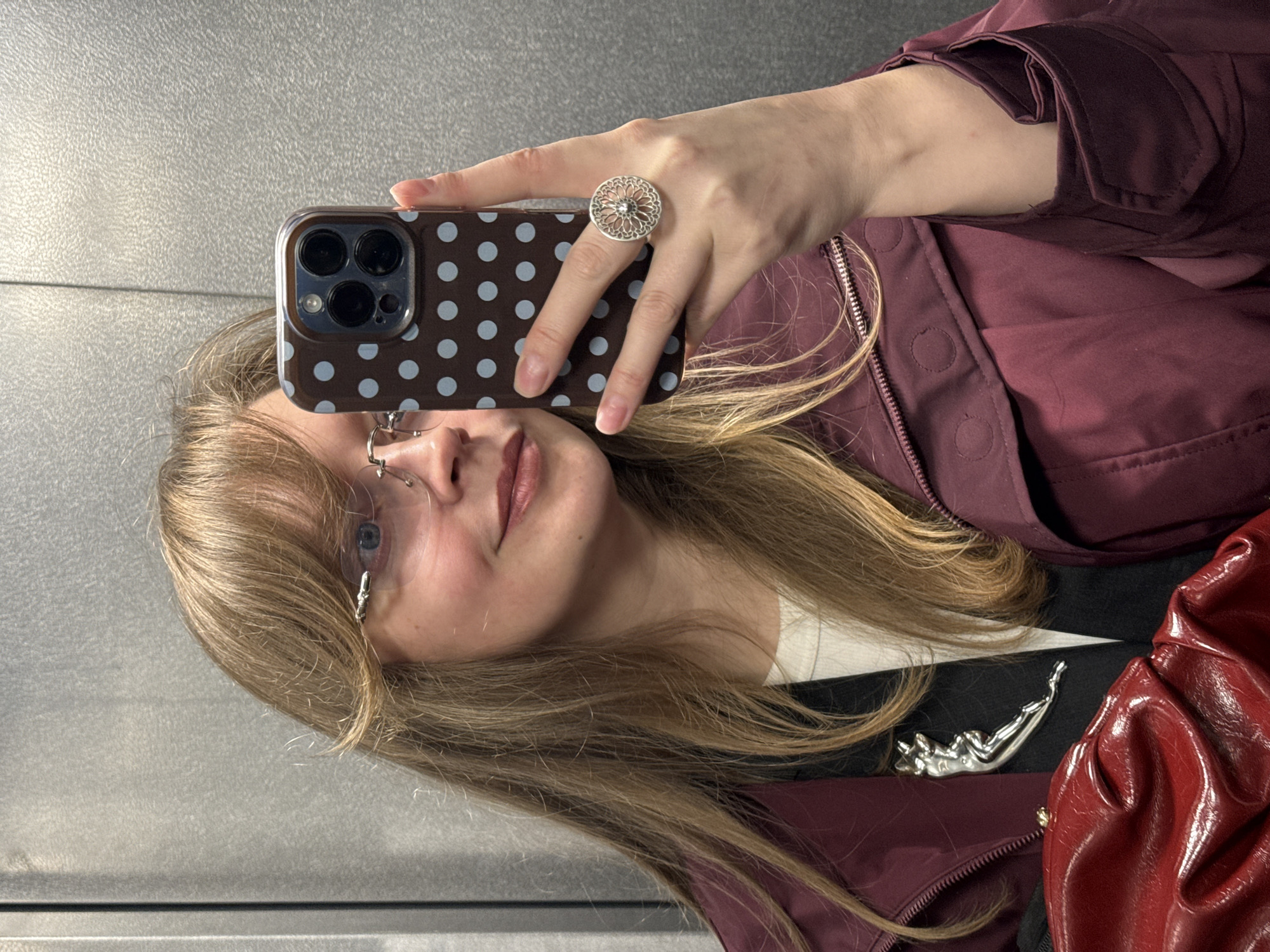 A selfie taken in an elevator. A young woman with blonde hair and glasses holds a phone with a brown polka-dot case, obscuring part of her face. She wears a large silver ring. The woman is wearing a burgundy jacket over a black and white top, adorned with a silver pin of a reclining figure. A shiny red bag is also visible. The background appears to be the metal interior of an elevator. The location is not discernible as Izhevsk, Russia, based on the visual elements provided.