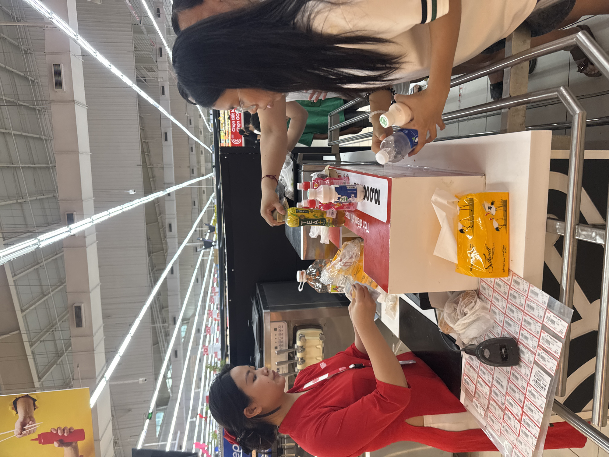 In Đông Hòa Lư Ward, Vietnam, a retail transaction is underway at a checkout counter in what appears to be a supermarket or convenience store. A female cashier, dressed in a traditional red Áo dài with white trim, stands behind the counter, holding what looks like small receipts. She is interacting with a female customer who is reaching for bottles from a display. The customer, wearing glasses and a white shirt with green and navy stripes on the sleeves, is holding two bottles, one clear and one opaque. Behind them, other shoppers are visible, including a person in a green shirt. The counter area is equipped with a soft-serve ice cream machine, a barcode scanner, and a box of tissues featuring cranes. The environment is brightly lit with fluorescent ceiling lights, typical of a commercial establishment, and the background shows shelves stocked with goods and store signage.