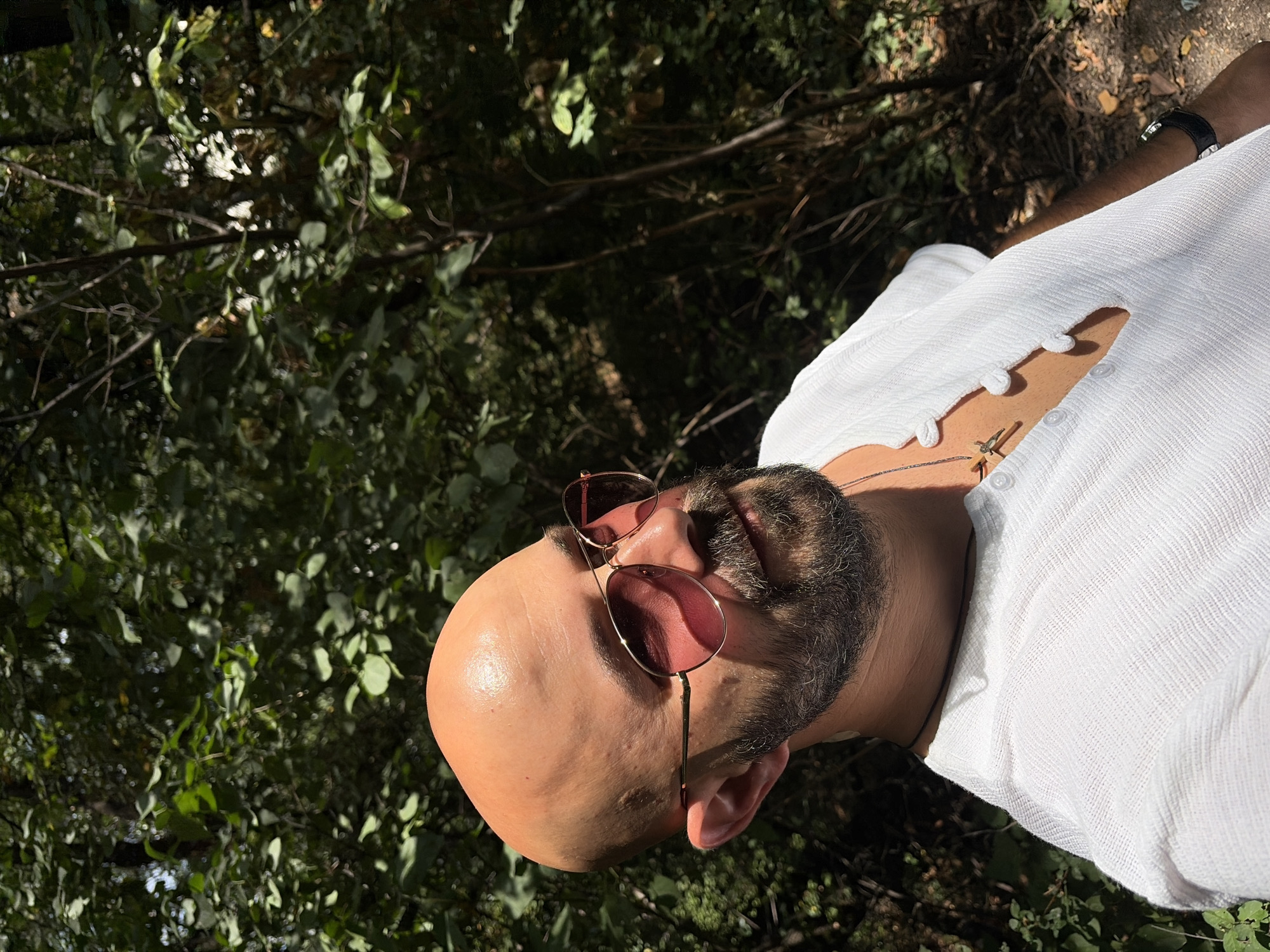 A bald, bearded man wearing sunglasses and a white shirt stands outdoors in a wooded area. Sunlight dapples through the leaves, creating a natural, shaded environment. The man's head is turned slightly upwards and to his left, with his gaze directed away from the camera. He has a goatee and a full beard. He is wearing aviator-style sunglasses with rose-tinted lenses. A silver cross necklace is visible around his neck, resting on the V-neck of his white shirt. The shirt appears to be made of a textured fabric and has a button-up placket with several visible buttons. A dark watch with a studded band is on his left wrist. The background consists of dense green foliage and tree trunks, suggesting a park or forest setting. The lighting suggests it is daytime. The location is identified as Sofia, Bulgaria.