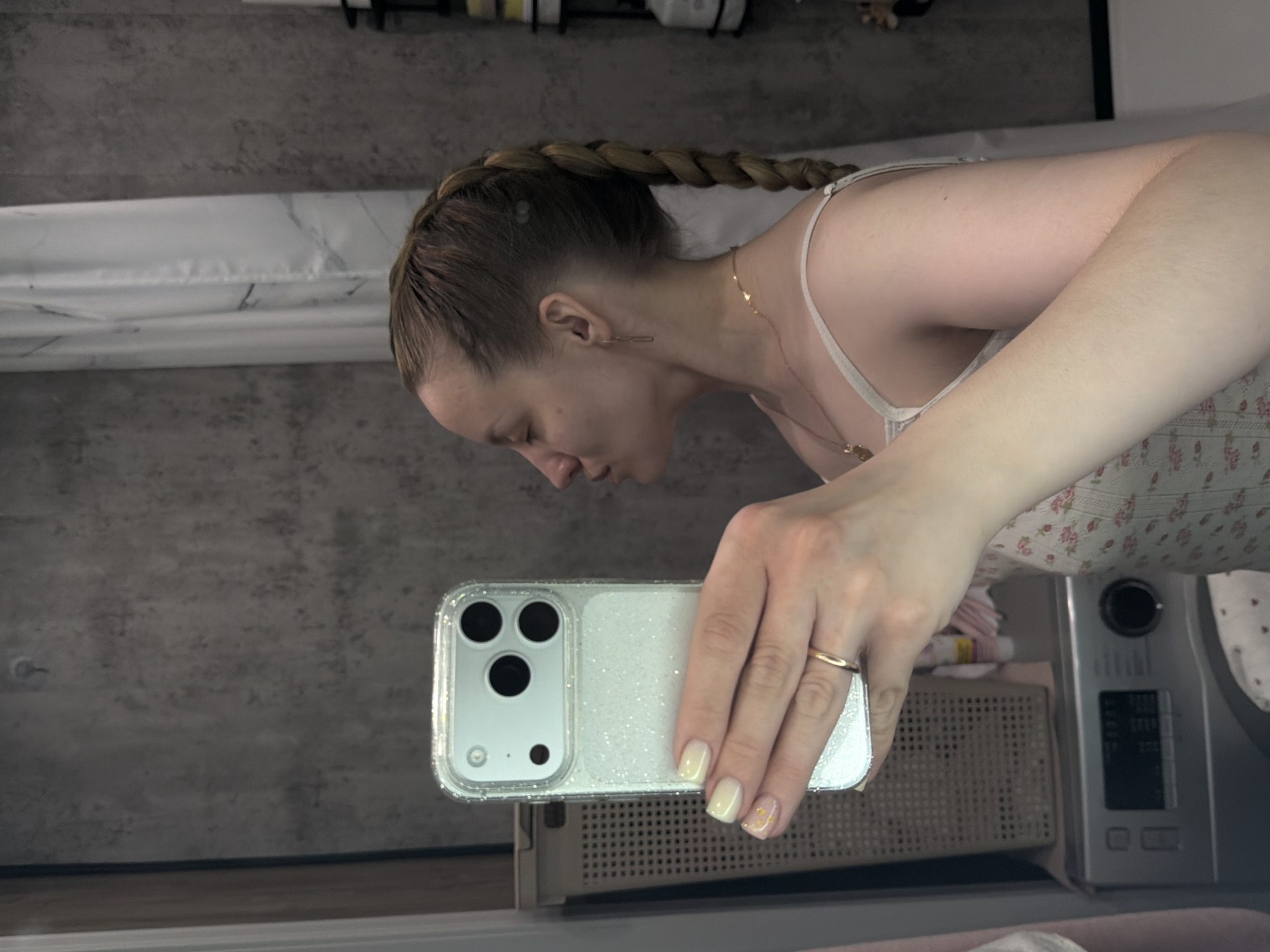 A woman with a braid is taking a selfie in a bathroom. She is holding a smartphone with a glittery case. The bathroom has a grey wall, a white marble bathtub, and a laundry machine. The location is not discernible from the image and therefore Reshetnikovo, Russia cannot be confirmed.