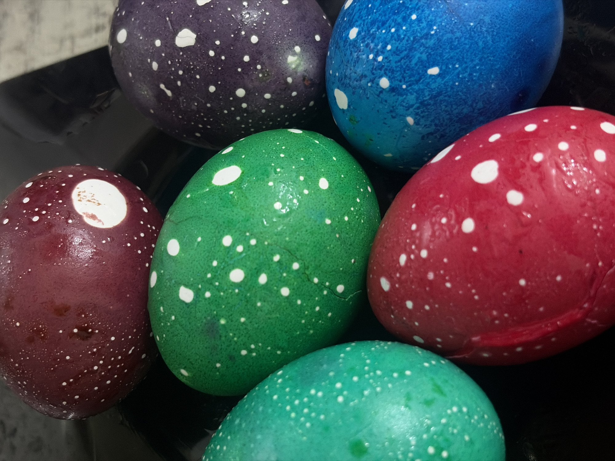 The image shows a close-up of six Easter eggs dyed in different colors: purple, blue, red, dark red, green, and turquoise. All of them have white speckles. They are lying on a black, possibly glossy, surface. The eggs are clustered together, partially overlapping each other. No specific details suggest a location in Saint Petersburg, Russia.