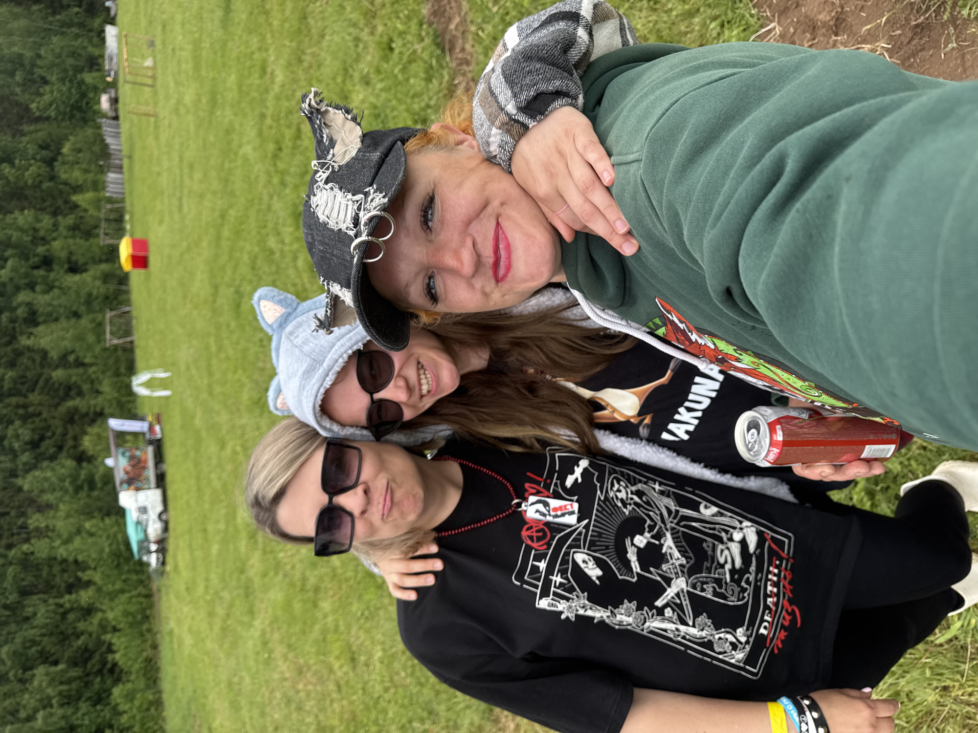 Three women are captured in a close-up selfie, embracing each other outdoors in a grassy field in Bol'shaya Kivara, Russia. The image shows them from the upper body, with the background consisting of green grass and trees.
The woman at the back has reddish-blonde hair, light makeup with red lipstick, and a broad smile. She wears a distressed black baseball cap adorned with safety pins and metal rings. Her attire consists of a dark green hooded sweatshirt layered over a grey and black plaid long-sleeved shirt. Her arm is around the woman in front of her.
In the middle, a woman with long brown hair wears a light blue hooded sweatshirt with ear-like features on the hood, and black sunglasses. She smiles and holds a red and white beverage can. Her black t-shirt has the text "AKUNA" visible on it.
The woman in the foreground has blonde hair and black sunglasses, with a subtle smile. She wears a black t-shirt featuring an intricate white graphic print, depicting skeletal figures and roses, consistent with gothic or metal aesthetics. The word "DEATH" is prominently displayed in red on her shirt. She accessorizes with a red beaded necklace and a white pendant. On her left wrist, she wears a yellow and blue bracelet alongside a black studded bracelet. Her arm is around the middle woman.
The setting is an open green field, with trees lining the distant background. Faintly visible are a red and yellow object, a wooden fence, and what appears to be a white vehicle or stall, suggesting an outdoor event or a casual gathering. The lighting indicates a daytime shot, possibly under an overcast sky, and the women's expressions convey happiness and camaraderie.