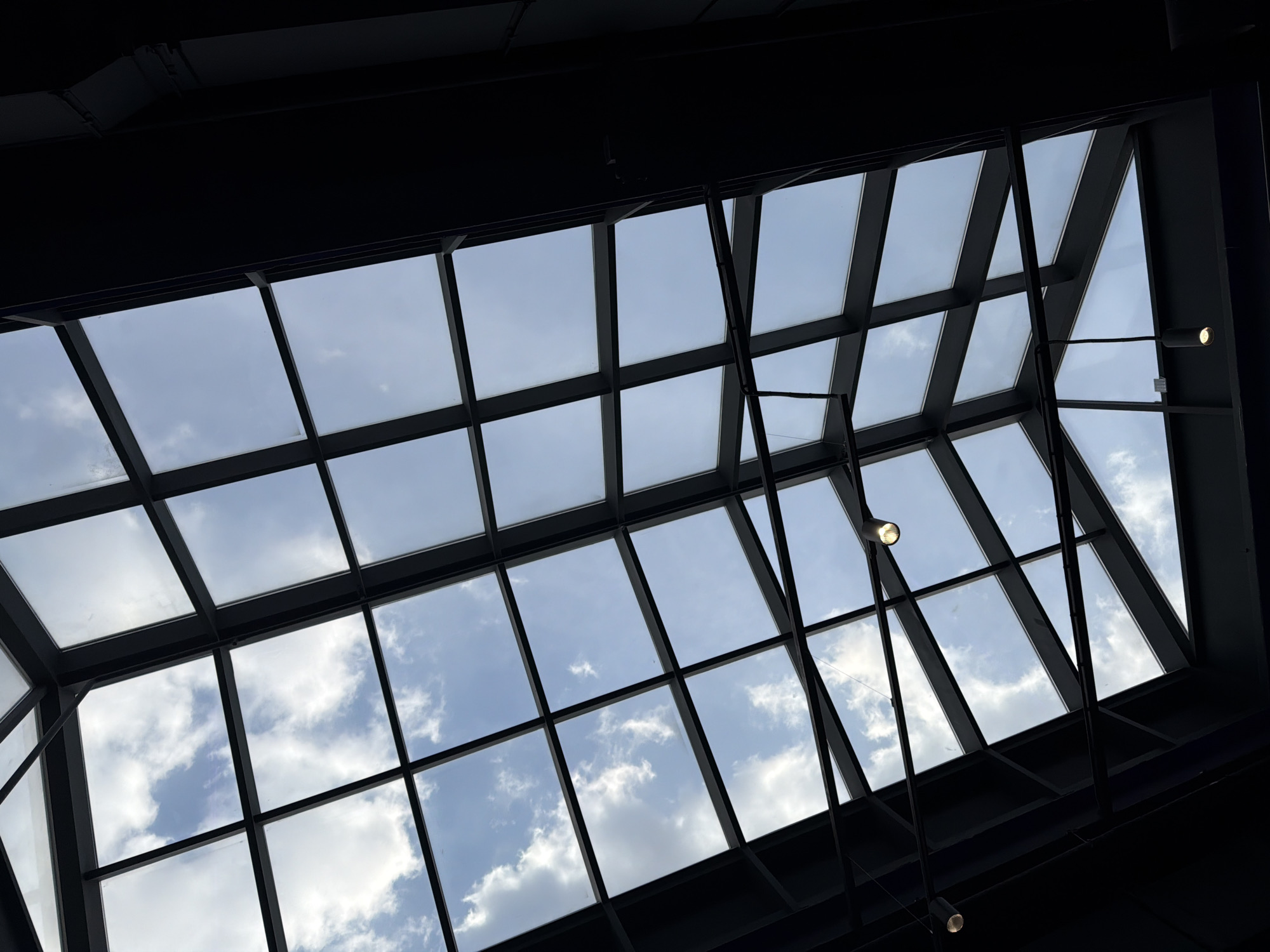 An upward-facing shot captures a large, gridded glass ceiling or skylight within a modern building. The dominant feature is the dark, angular metal framework that divides the transparent roof into numerous rectangular and trapezoidal panes. Through these glass panels, a bright, partially cloudy sky is visible, featuring scattered white cumulus clouds against a light blue background.
The structural framework creates a strong geometric pattern across the image. Dark, solid ceiling elements, likely supporting beams or the outer perimeter of the roof, frame the skylight on the top, bottom, and right sides, contrasting sharply with the bright sky. Two artificial light fixtures with illuminated bulbs are discernible: one positioned centrally on the right edge of the grid, and another smaller one lower down on the right.
The scene depicts an interior architectural view during daytime, characterized by the natural light filtering through the overhead glazing and the clear distinction between the man-made structure and the natural sky. The setting is in Krasnoyarsk, Russia. No people or visible text are present.