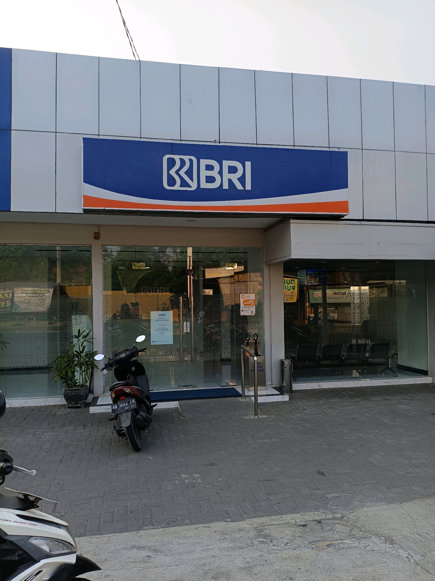 The image shows the exterior of a BRI (Bank Rakyat Indonesia) branch in Purwomartani, Indonesia. The bank has a modern facade with a large blue and orange sign displaying the BRI logo. The entrance features large glass doors, and the interior is visible, showing a waiting area with chairs. Outside, a black motorcycle is parked in front of the entrance, and another white and black motorcycle is partially visible in the foreground. The ground is paved with interlocking bricks. The sky is overcast, suggesting it is either early morning or late afternoon. The overall scene depicts a typical banking facility in a semi-urban Indonesian setting.