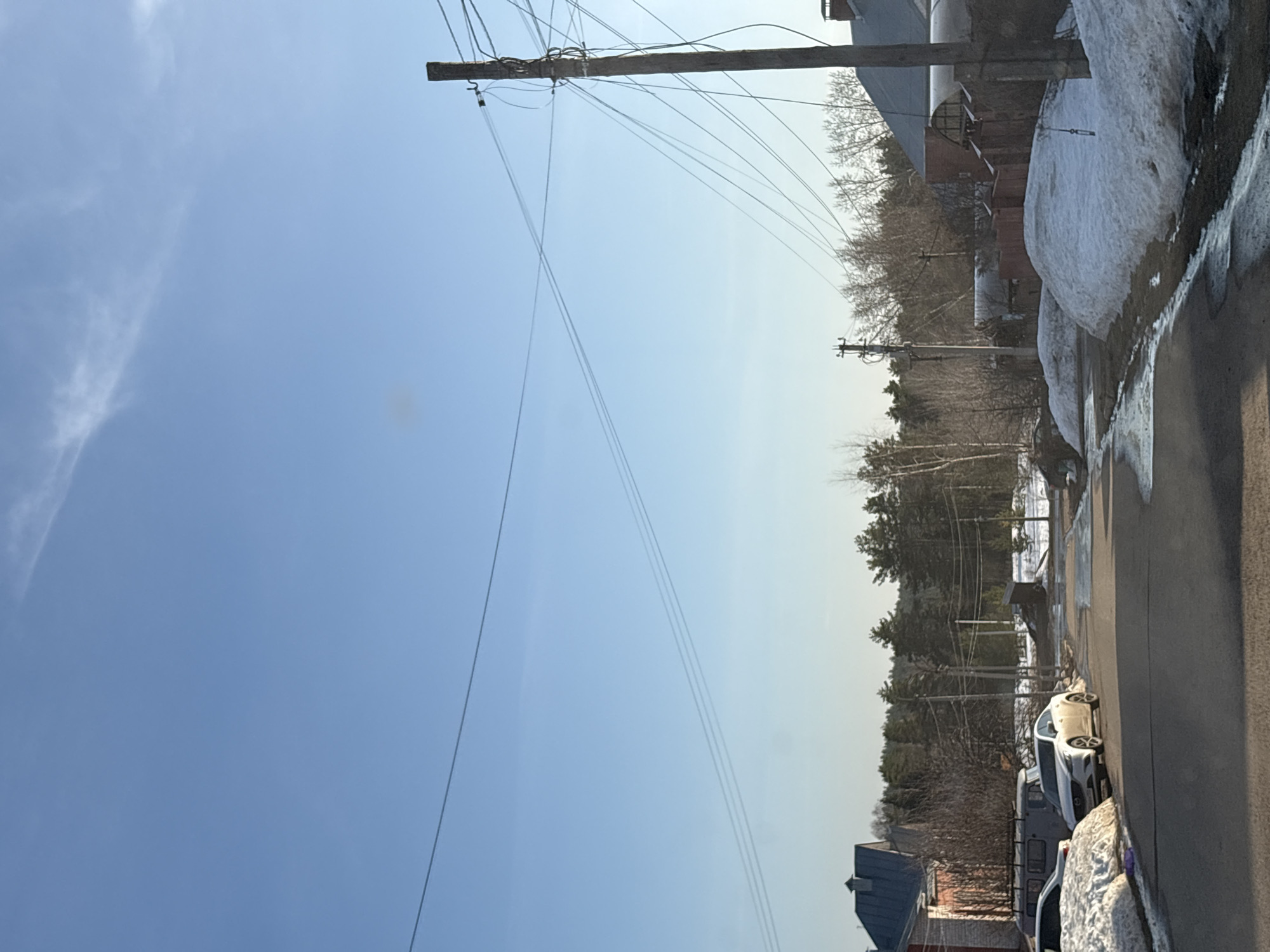 The image shows a street scene in Прикамский, Russia, on a day with clear blue sky and some clouds. Utility poles and overhead power lines are visible. Snow piles line the side of the street, and several cars are parked along the road. In the background are trees and buildings, including what appears to be residential structures. The road is paved.