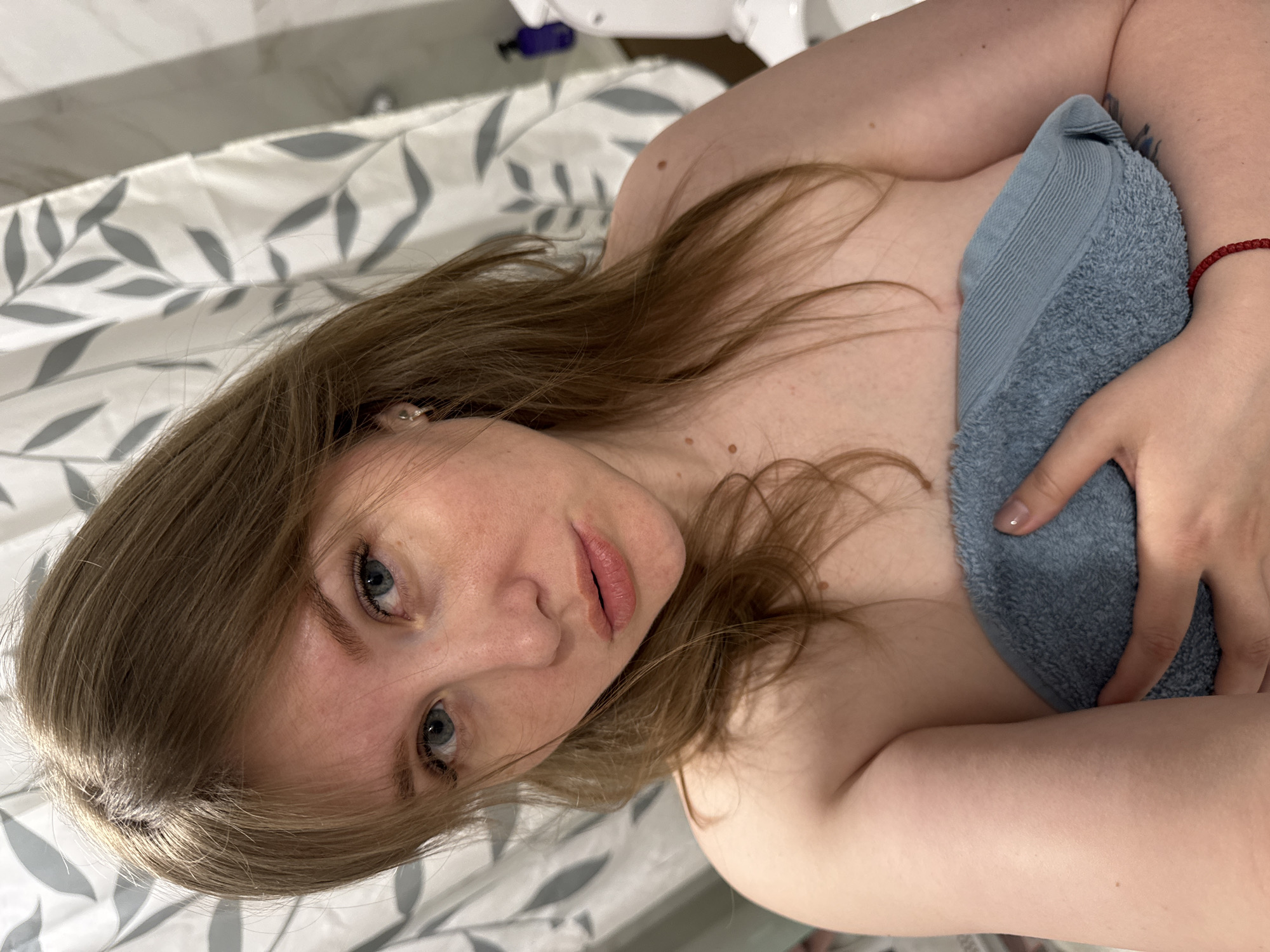 A woman with light brown hair and blue eyes is lying down, holding a light blue towel. The background appears to be a bathroom with a shower curtain patterned with gray leaves. The location is not identifiable as Gornyi (Dvurechenskoe S/P), Russia, based on the provided image.
