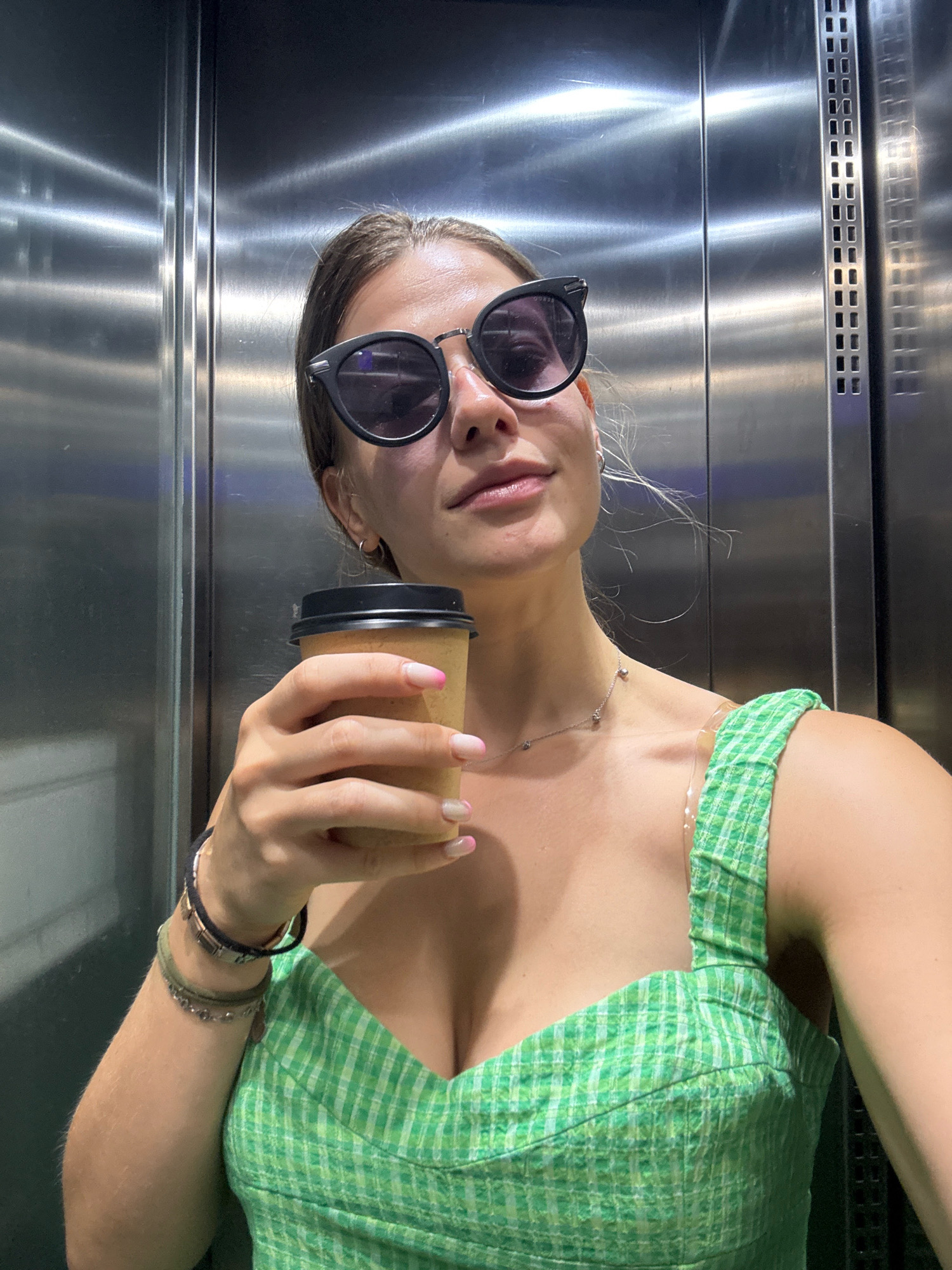 A young woman with brown hair, wearing large black sunglasses and a green plaid top, stands in what appears to be an elevator. She is holding a disposable coffee cup in her right hand, which is raised to chest level. The elevator walls are made of brushed metal, reflecting the overhead lighting. Her left arm is adorned with several bracelets. The overall impression is of a casual, everyday moment captured within an urban environment. The specific location within the city of Barcelona, Spain, cannot be determined from the visual information present in the image.