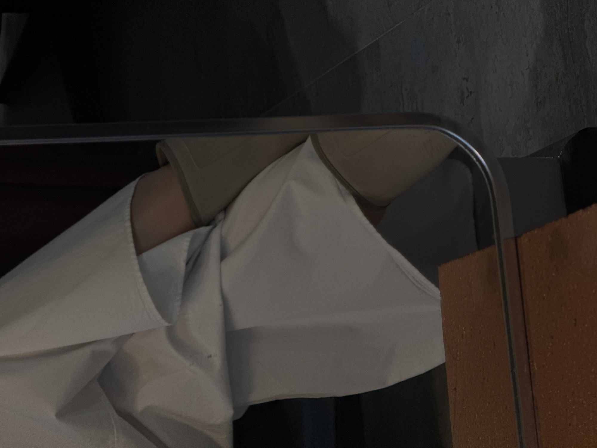 The image shows a partial view of a tanning bed, with a white sheet partially covering the surface. The headrest is visible under the sheet. The background features a textured wall with a grey hue. A brick or tile element is present on the right side of the image. The location cannot be determined to be Yerevan, Armenia, based on the provided image.