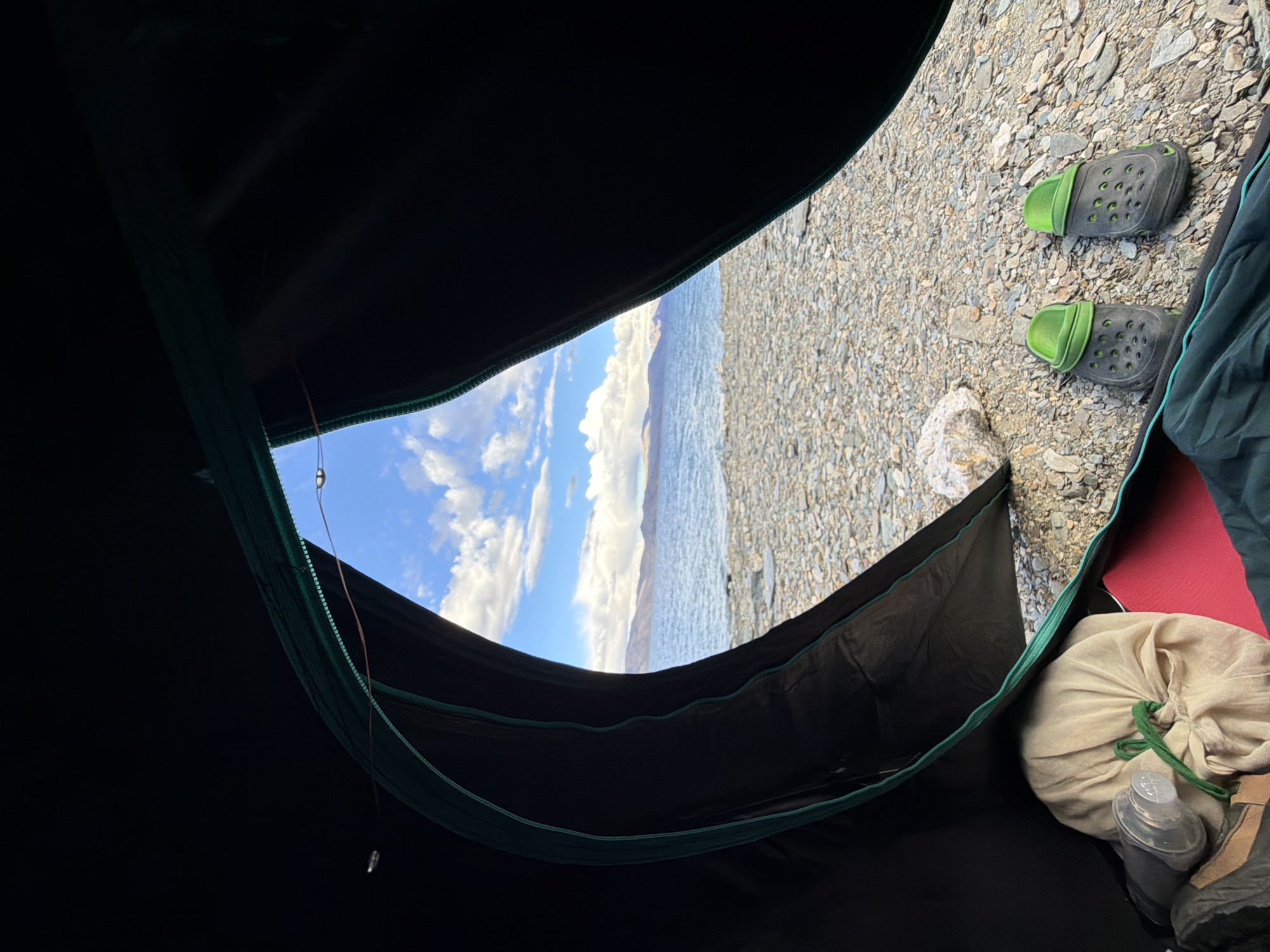 The image shows the view from inside a dark tent, looking out at a rocky shoreline and a large body of water under a partly cloudy sky. In the foreground, inside the tent, a beige drawstring bag and a clear plastic container are visible. A red mat and part of a green sleeping bag are also present. Outside the tent, on the rocky ground, are a pair of green and grey clogs. The water is calm, and mountains are visible in the distance. The scene suggests a camping location by a lake in a remote, mountainous area. The location is likely in Ladakh, India, known for its high-altitude lakes and stark landscapes.