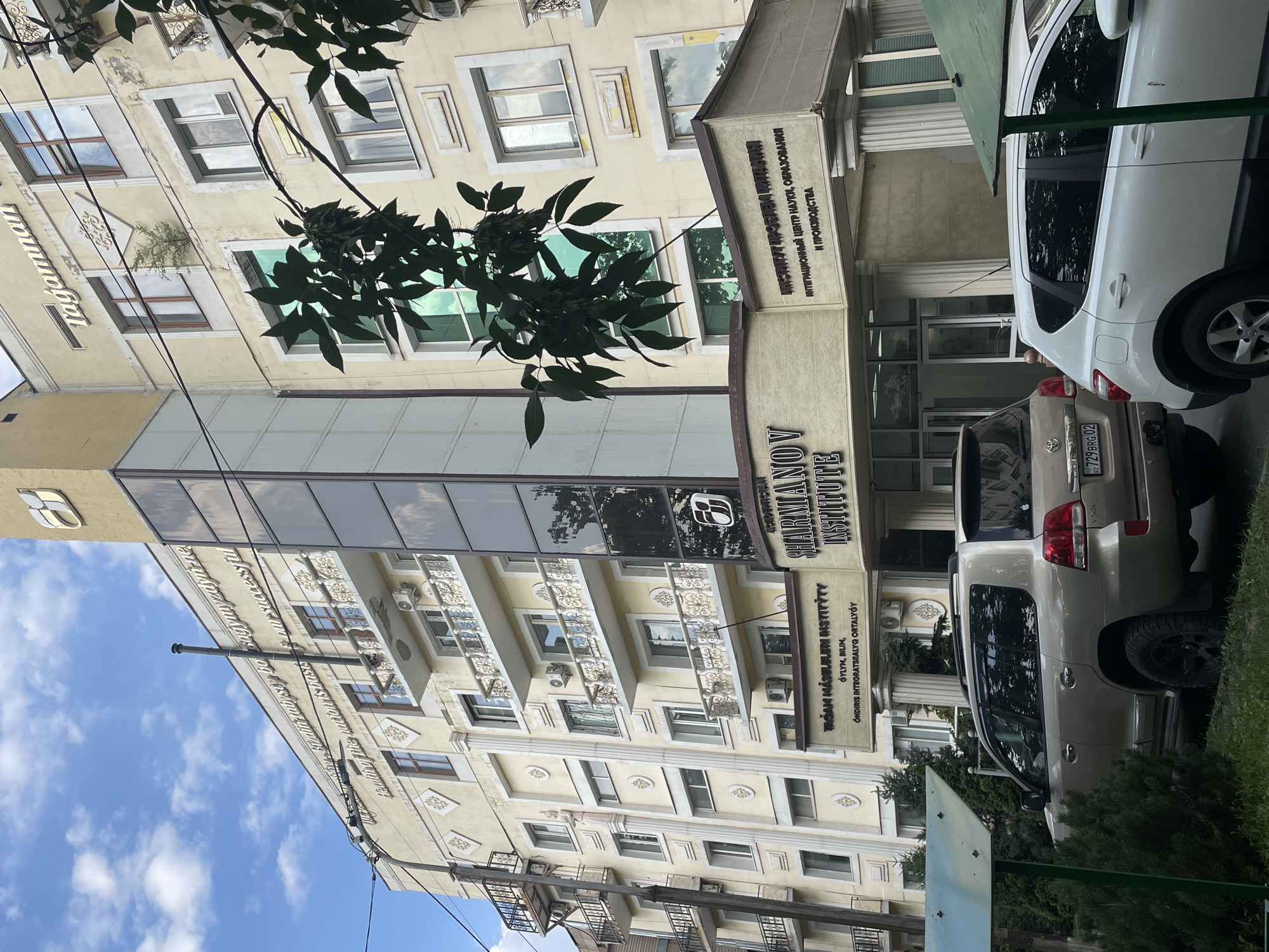 The image shows the Sharmanov Institute in Almaty, Kazakhstan. The building is a pale yellow, multi-story structure with a glass elevator shaft running up the side. Above the entrance, signs in Cyrillic and Latin script identify the building as the "Sharmanov Institute" and "INSTITUT PROBLEM OCHISCHENIYA INTEGRATSIONNYY TSENTR NAUKI, OBRAZOVANIYA I PROIZVODSTVA". Another sign on the building reads "Tagamnan". A number of cars, including a beige SUV and a white hatchback, are parked in front of the building. The sky is blue with scattered clouds. Lush green trees and bushes surround the building, with a tree branch obscuring part of the upper floors. The architecture suggests a modern building with some classical influences.