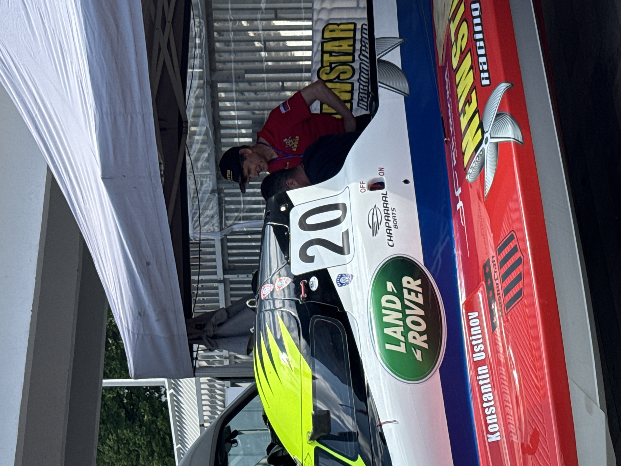 The image shows a close-up view of a racing boat, likely at a sporting event. The boat is white with red and blue accents, and features several sponsor logos, including "LAND ROVER" and "NEW STAR racing team". The number "20" is prominently displayed on the side of the boat. Two men are visible near the boat; one, wearing a red shirt and a baseball cap, appears to be interacting with the other man who is partially obscured. The scene is partially covered by a white tent, suggesting an outdoor event. The location is identified as the city of Perm, Russia.