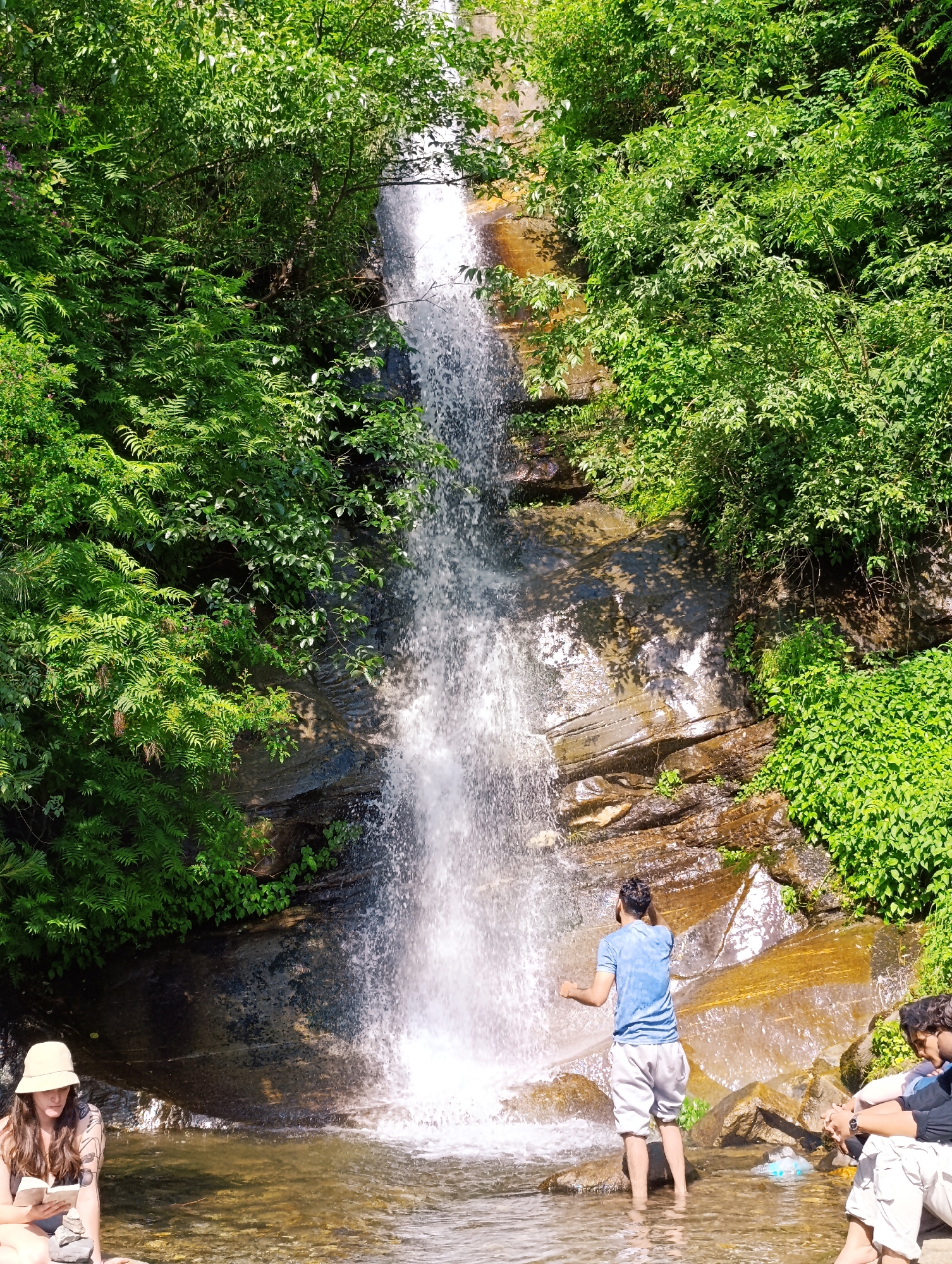 In Vashisht, India, a picturesque scene unfolds near a cascading waterfall. A woman, wearing a light-colored bucket hat, sits on a rock by the water's edge, engrossed in reading a book. To her left, a stack of small stones rests on the rock. The waterfall plunges down a rocky cliff face, surrounded by lush green foliage and trees. In the middle of the frame, a young man in a blue t-shirt and white shorts stands in the shallow water, with his back to the camera, his arms outstretched towards the waterfall. To the right of the frame, another person, partially visible, sits near the water. The environment is vibrant with natural colors, featuring the white of the falling water, the earthy tones of the rocks, and the rich greens of the vegetation under a bright, sunny sky.