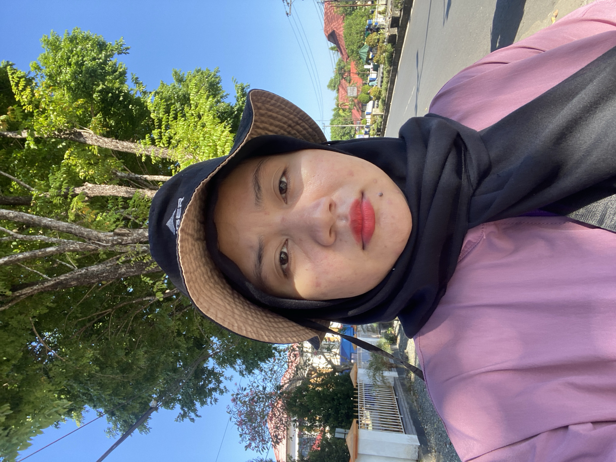 A young woman wearing a black hijab, a pink jacket, and a black bucket hat is taking a selfie outdoors on a sunny day in the city of Banjarbaru, Indonesia. She is standing on a sidewalk next to a road with a fence and some houses visible in the background, as well as trees and a clear blue sky. The woman has red lipstick on and appears to be looking directly at the camera.