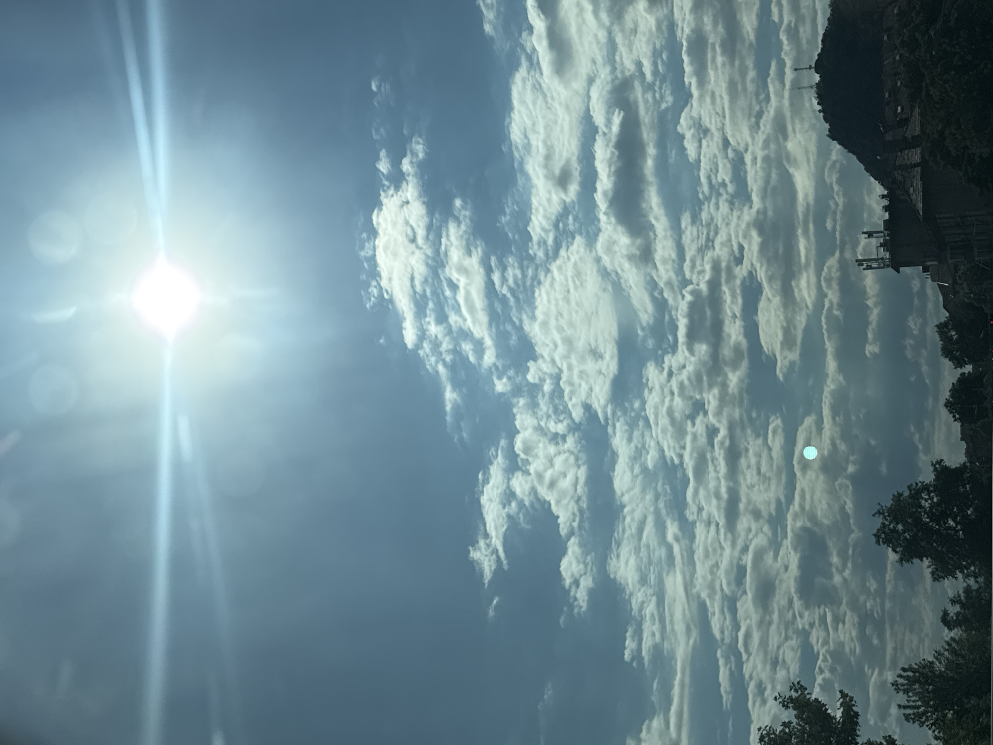 The image captures a sky filled with white, fluffy clouds under a bright, hazy sun, creating a lens flare effect. In the lower portion of the image, a silhouette of a hilly landscape is visible. On the hills, there are structures that appear to be buildings, and on the highest peak, communication towers are discernible. The lower foreground shows the dark silhouettes of trees and building rooftops. The scene is consistent with a view of the hills of Plovdiv, Bulgaria, perhaps the Nebet Tepe or Dzhambaz Tepe area, given the presence of historical structures and modern communication towers on the hills. The lighting suggests it is daytime, likely mid-morning or afternoon, with the sun positioned high in the sky. There are no people or animals visible in the frame.