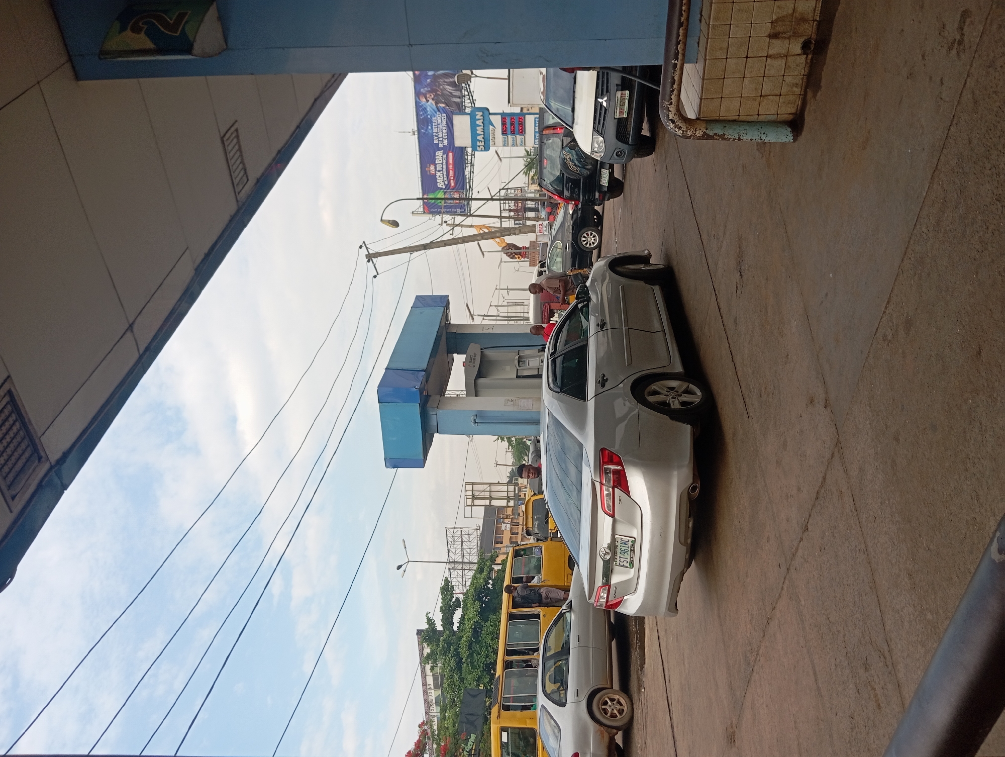 The image shows a view of a gas station in Onitsha, Nigeria. Multiple vehicles, including cars and yellow buses, are present. The price of gas is visible on a sign with the word SEAMAN prominently displayed above. The backdrop includes a billboard, power lines, and buildings. Some people are standing near the gas pumps. The sky is visible with a few clouds.