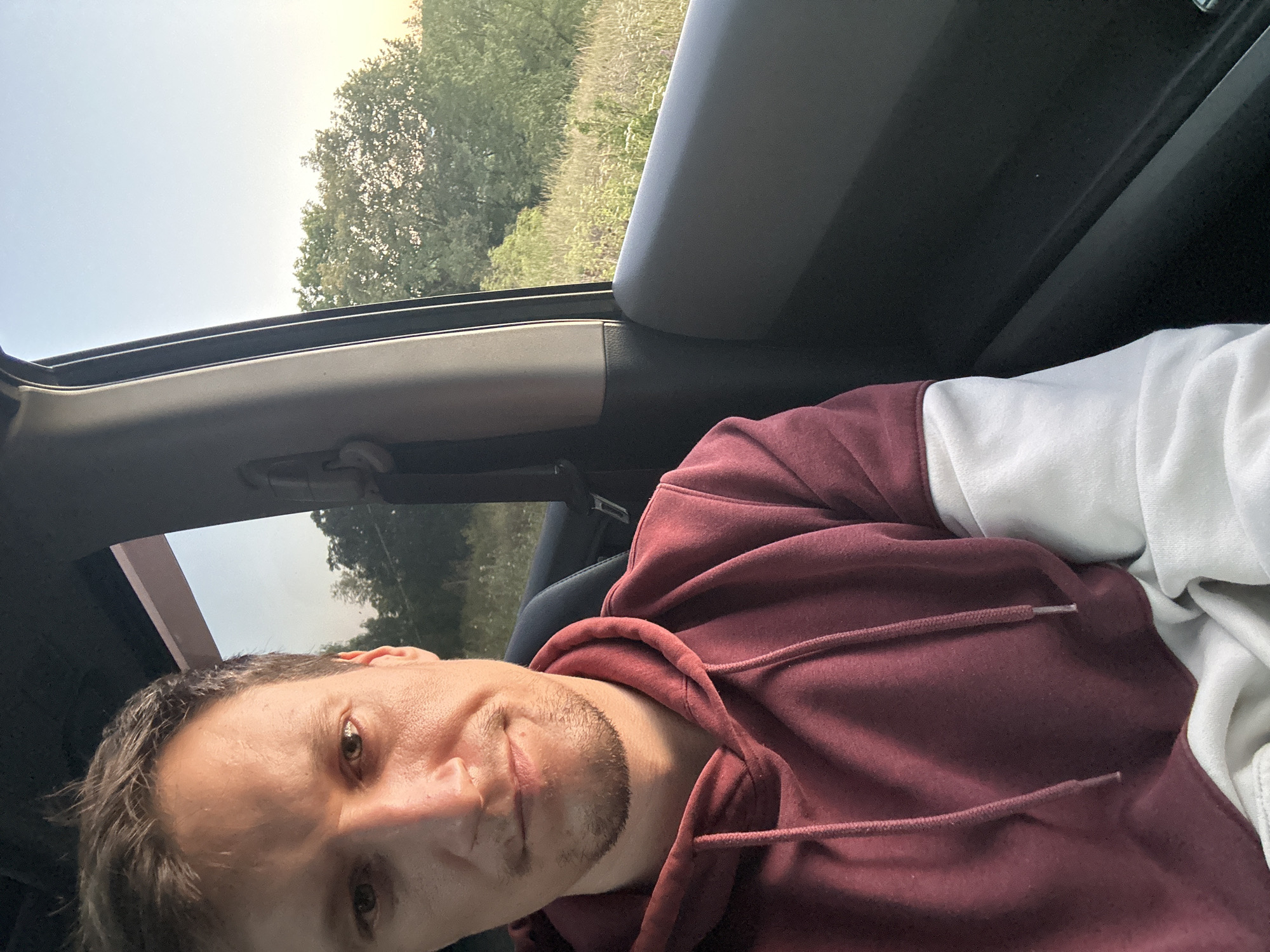A selfie taken from inside a vehicle shows a Caucasian male looking towards the camera. He has short brown hair, a light complexion, and is wearing a maroon hooded sweatshirt with white sleeves. The man has a slight smile and appears to be in a rural or natural setting, as indicated by the trees and vegetation visible through the car window. The sky in the background suggests it may be late afternoon or early evening. The interior of the vehicle is dark, with a portion of the car door visible.