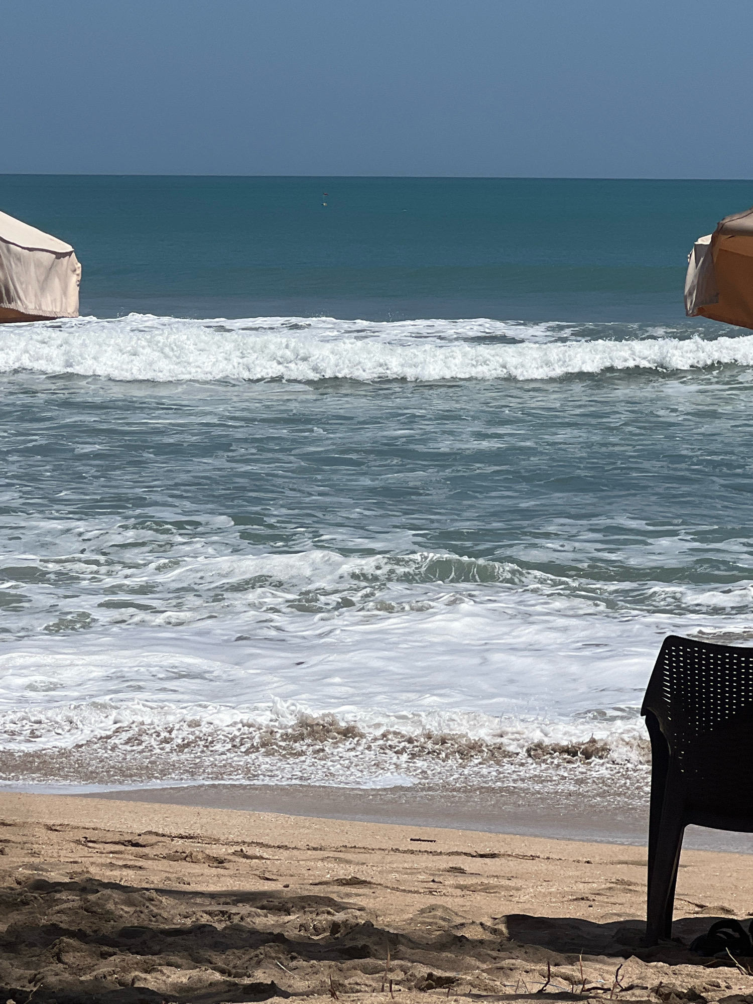 This image captures a serene beach scene in Kuta, Indonesia. The ocean is a prominent feature, with gentle waves rolling onto the sandy shore under a clear blue sky. Two beach umbrellas, one on the left and one on the right, provide shade on the beach. A black plastic chair is positioned in the foreground, facing the ocean, suggesting a relaxing atmosphere. The sand is textured with footprints and some debris, indicating recent activity on the beach. In the distance, two small buoys are visible in the water, adding a subtle detail to the vastness of the ocean. The overall impression is one of tranquility and natural beauty, typical of a tropical beach setting.