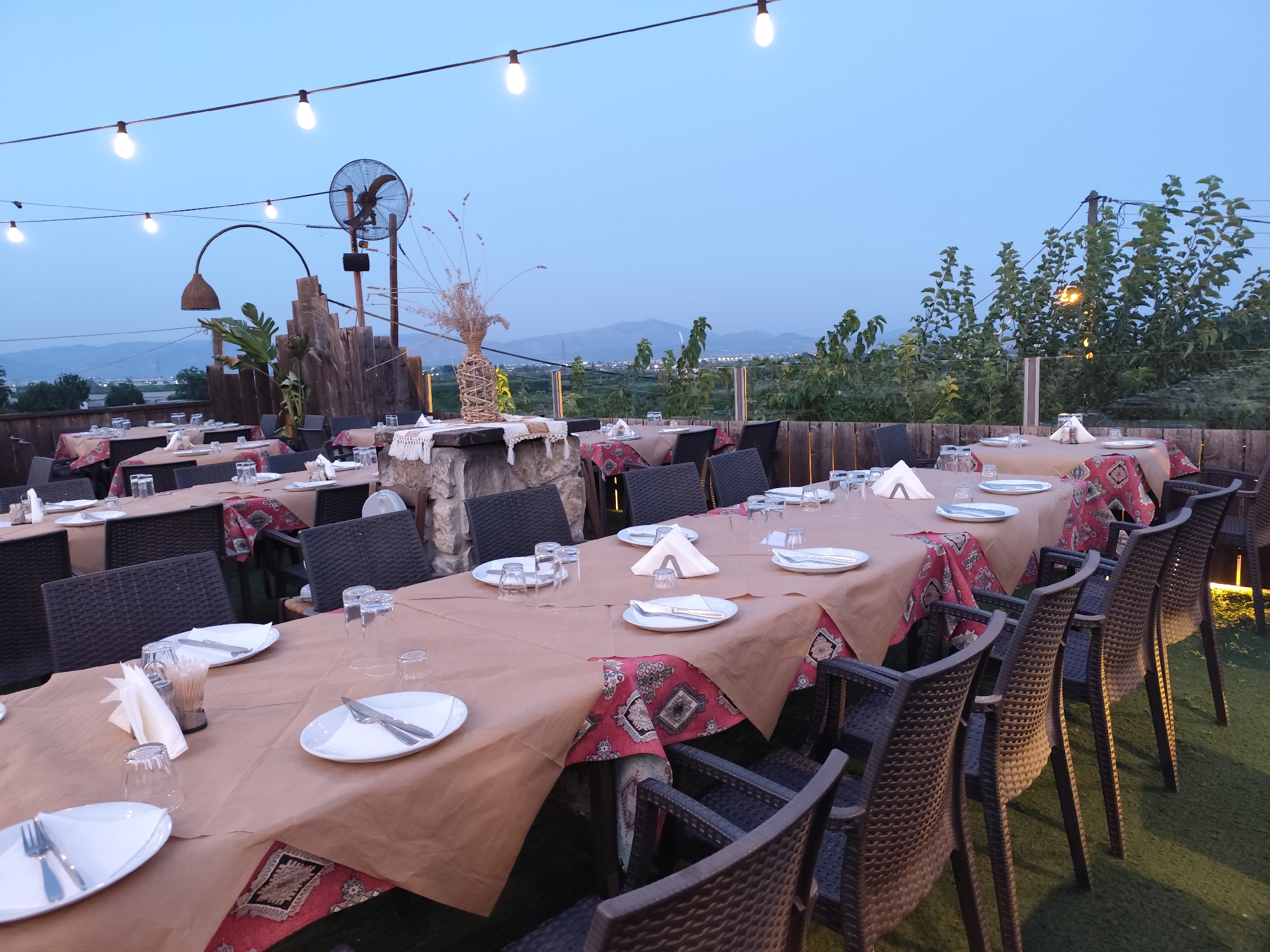 An outdoor dining area is set up with multiple tables covered in brown paper and patterned tablecloths, arranged with place settings including white plates, silverware, and glasses. Dark brown wicker chairs surround the tables. String lights are suspended overhead, and a large industrial fan is mounted on a post in the background. The scene overlooks a landscape with trees and distant hills, with the sky a dusky blue. It appears to be located on a rooftop or elevated patio within the Municipal Unit of Argos, Greece, possibly a restaurant or event space given the setup.