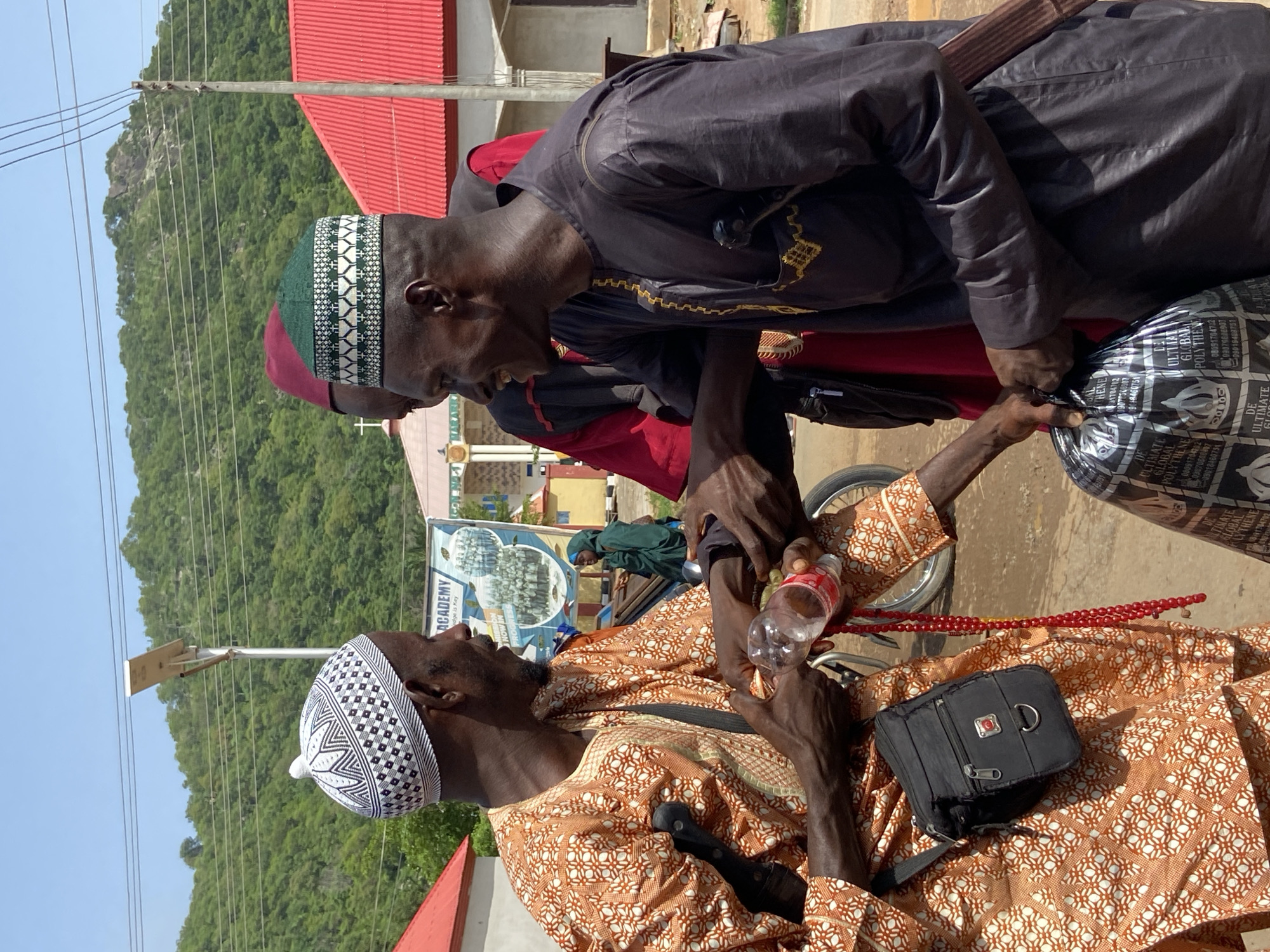 The image captures a street scene in Mararaba, Nigeria. Three men are the central figures. One man is wearing an orange and white patterned robe and a white patterned cap. He is holding a water bottle and prayer beads. Another man is dressed in a black robe and a green patterned cap, with hints of red clothing underneath. The third man is partially visible behind the man in the black robe. All three men appear to be engaged in a conversation, possibly exchanging or examining items.
In the background, there is a building with a red roof, along with a sign that includes the word "ACADEMY". Beyond the building, a landscape with greenery and a hill or mountain is visible. Utility poles and wires stretch across the sky. The setting suggests a bright, daytime scene with a mix of urban and natural elements.