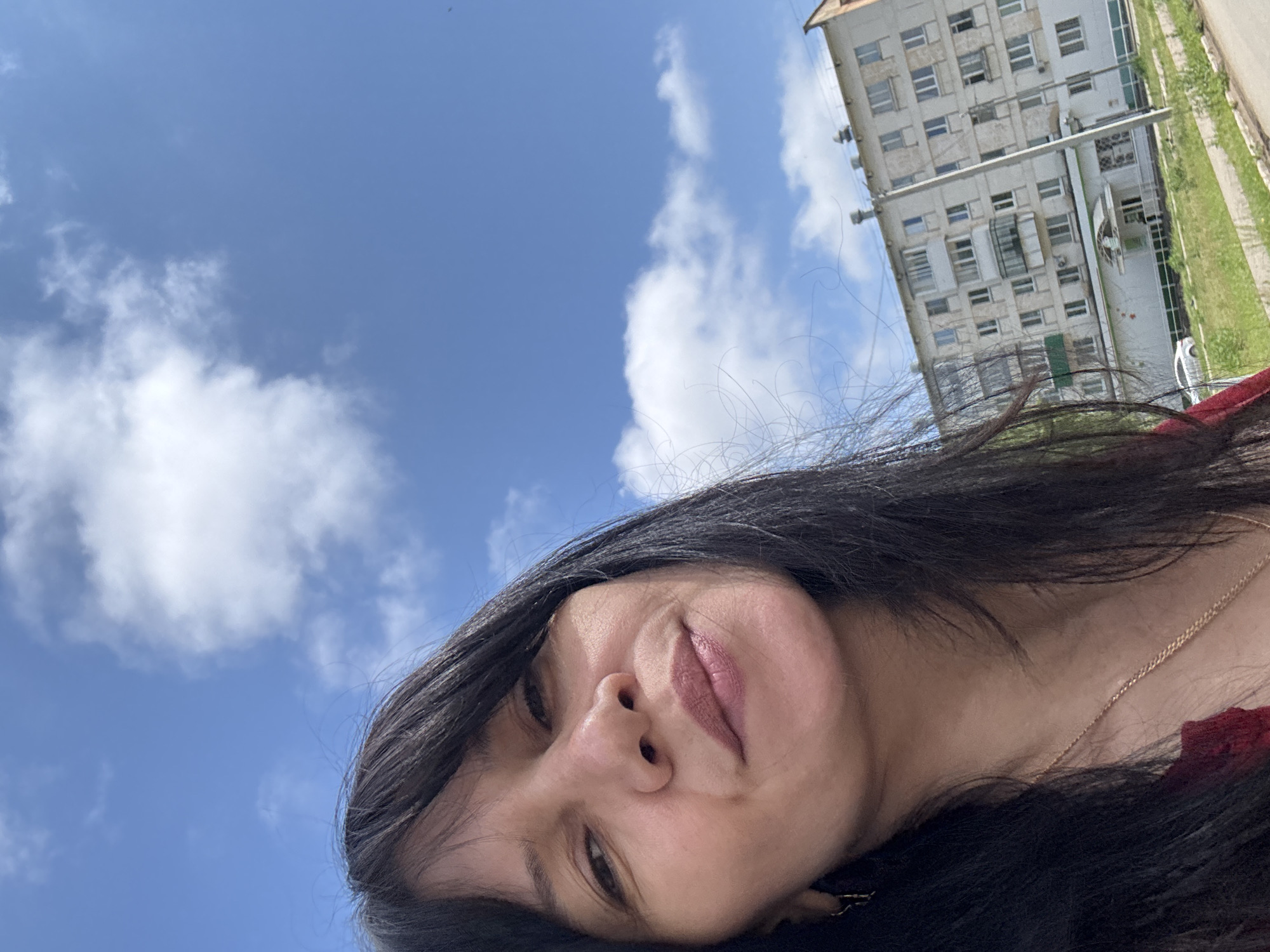 The image is a brightly lit, upward-angled selfie featuring a woman against a backdrop of a clear blue sky, scattered white clouds, and a multi-story apartment building. The photograph is rotated clockwise, causing the woman's head to be tilted and the background building to appear on its side.
The primary subject is a woman, likely an adult, whose face occupies the lower right portion of the frame. She has long, dark hair, some strands of which are visibly backlit and flowing across the top of the image. Her lips have a subtle, soft pink shade, and she exhibits a calm, slightly smiling expression while looking towards the camera. A delicate gold-colored chain necklace is visible around her neck.
In the background, a large, light-colored apartment building stands tall, characterized by numerous windows and balconies. Some parts of the building show signs of wear or age. Utility poles or lines are visible in front of the structure. To the right of the building, a strip of green grass or vegetation indicates an outdoor, possibly urban or suburban, setting.
The time of day appears to be sunny daytime with bright overhead light, suggesting clear weather conditions. The location context indicates this scene is in Chaikovsky, Russia. No visible text is present in the image.