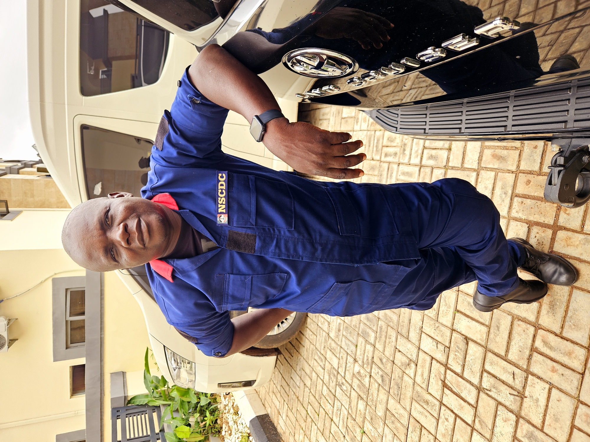 A bald, dark-skinned man wearing a blue jumpsuit with a red collar and a black undershirt stands leaning against a black vehicle. He has a patch on his left chest that says "NSCDC." A black smartwatch is visible on his left wrist. The man is standing on a light-colored, brick-patterned paved surface. In the background, a white van is parked. Behind the van, there is a light yellow building with grey trim and some windows. Green plants are visible to the left of the man and the van. The scene appears to be outdoors in a paved area, possibly a parking lot or driveway. The overall lighting suggests it is daytime, with overcast skies.