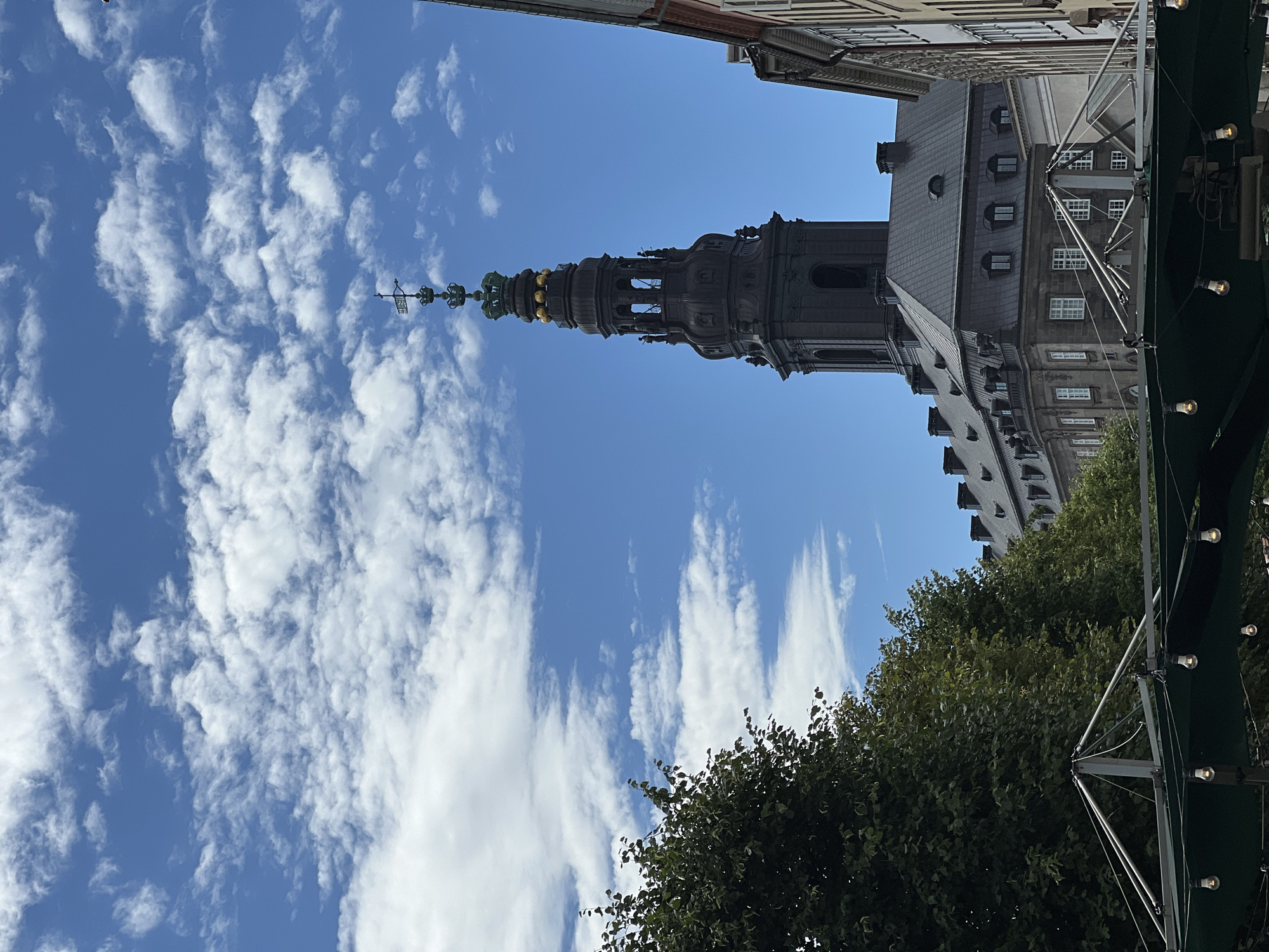 The image depicts a view of the Church of Our Lady (Vor Frue Kirke) tower in Copenhagen, Denmark. The dark, ornate tower, topped with crowns and a weather vane, rises against a vibrant blue sky filled with scattered white clouds. Below the tower, the dark-tiled roof of a building is visible, with dormer windows. To the left, lush green trees partially obscure the view, and in the foreground, a dark green awning with illuminated bulbs hangs, suggesting an outdoor seating area or market stall. The architectural style of the buildings suggests a historic European city.