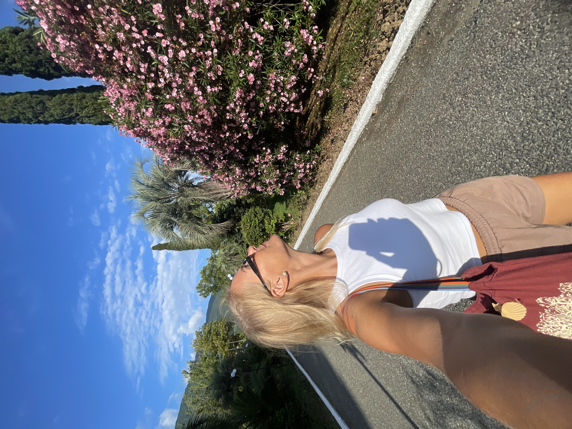 A blonde woman in sunglasses and a white tank top is taking a selfie while walking on a paved path. She is wearing brown shorts and carrying a maroon tote bag. Behind her, a large bush of pink flowers is in bloom, with two tall cypress trees and a palm tree visible. The background shows a lush green landscape with rolling hills under a blue sky with scattered clouds. The scene appears to be in a park or garden area within the city of Gagra, Abkhazia.
