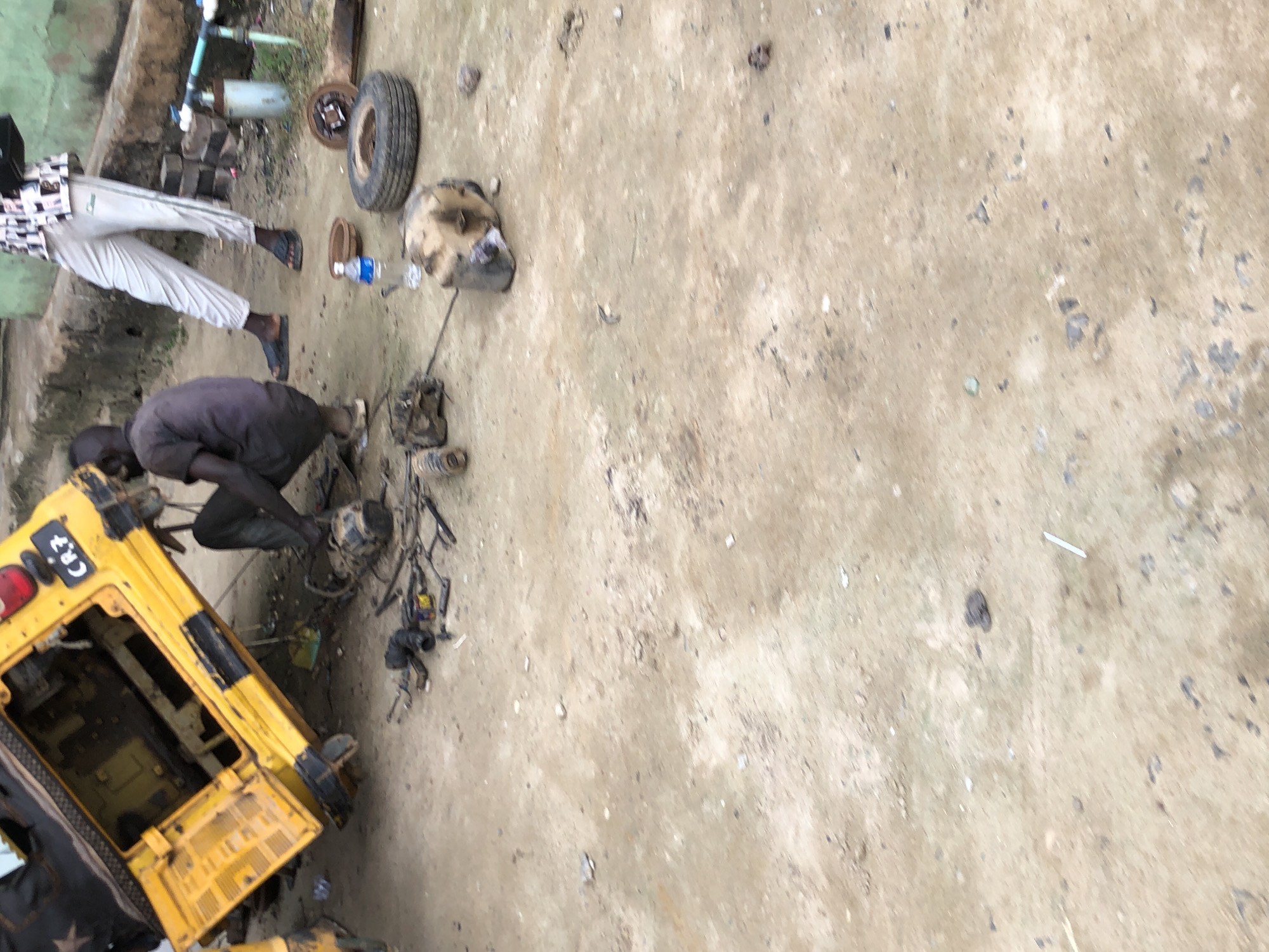 The image depicts an outdoor scene in Lagos, Nigeria, with a yellow auto-rickshaw, commonly known as a "Keke," tilted on its side. A person, appearing to be a male, is crouched beside the Keke, seemingly engaged in repairs or maintenance. Scattered around the Keke are various mechanical parts, tools, and a discarded tire, suggesting an ongoing repair operation. Another individual, wearing white pants and a patterned shirt, stands in the background, observing the scene. The ground is composed of dirt and gravel, indicative of a rough, unpaved area, typical of some residential or commercial spaces in Lagos. The overall environment appears to be a makeshift workshop or a roadside repair spot.