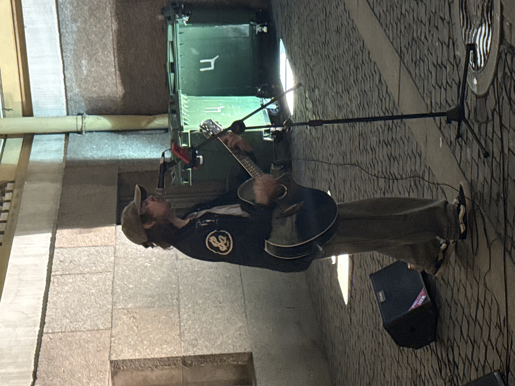 A person wearing a baseball cap and a dark jacket with a circular emblem on the left side, plays an acoustic guitar. The person is standing on a cobblestone surface in what appears to be a city square or street. A microphone on a stand is positioned in front of them, and a small black speaker is on the ground to their left. In the background, two large green dumpsters are visible, one with a Cyrillic letter "Ц" on it. The overall lighting suggests it is daytime, with some shadows cast on the ground. This scene is taking place in Saint Petersburg, Russia.