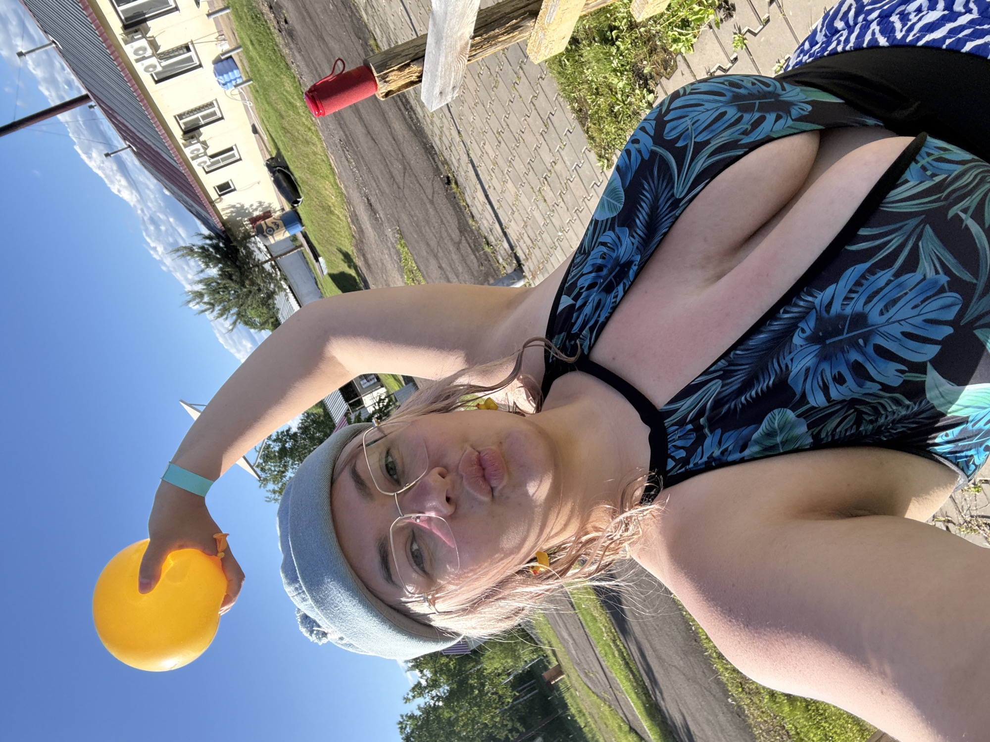 A woman is taking a selfie outdoors on a sunny day, holding a yellow balloon in her raised left hand. She is wearing a light blue knitted hat, glasses, and a black and blue tropical-print swimsuit. Her lips are pursed, and she has yellow duck-shaped earrings. In the background, there are buildings with red roofs and white walls, trees, a paved area, and grass. A red speaker is mounted on a wooden post. The image appears to be taken in an outdoor recreational area.