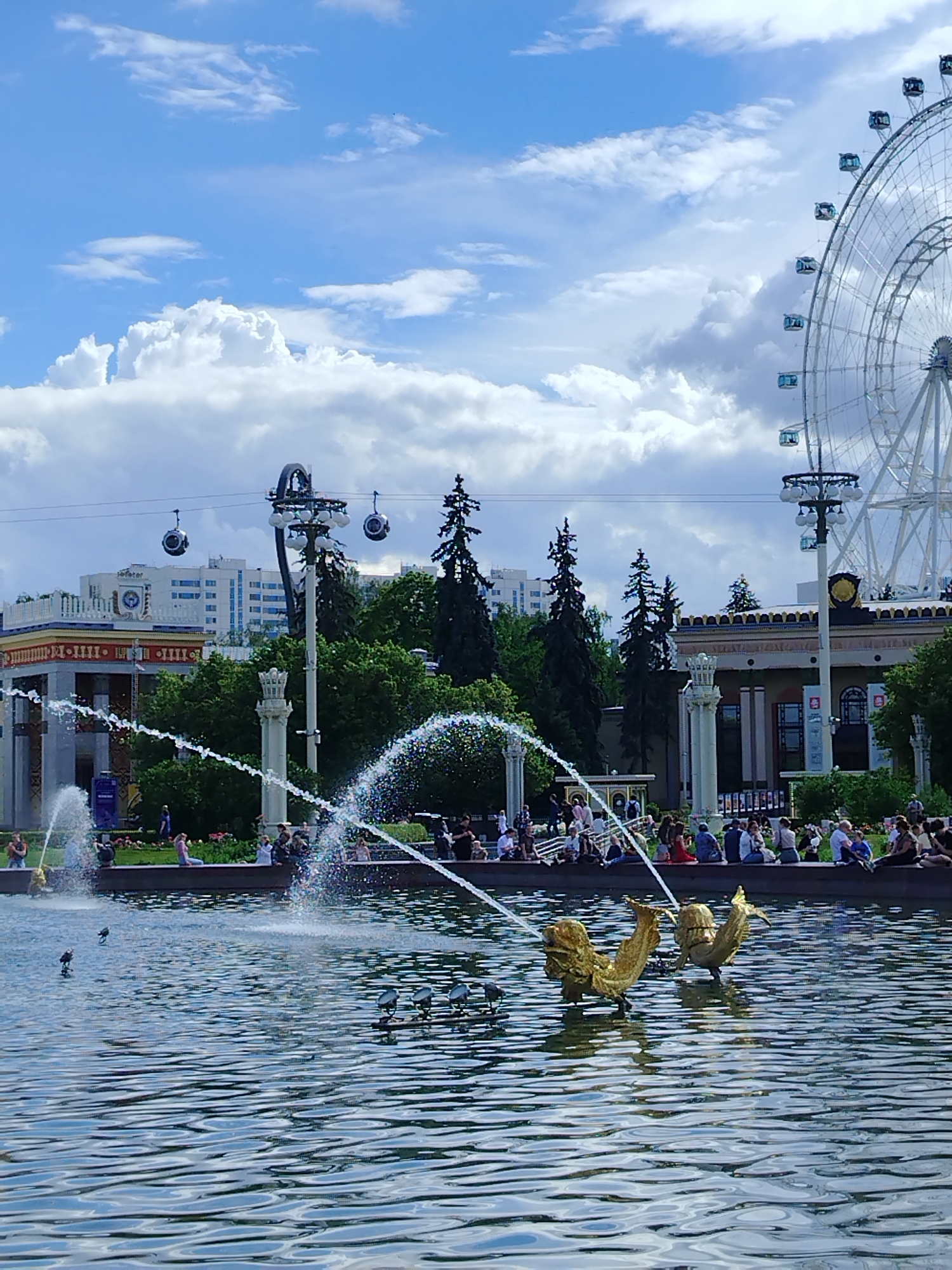 The image depicts a bright, sunny day in Moscow, Russia, with a clear blue sky dotted with fluffy white clouds. In the foreground, a tranquil body of water reflects the sky and surrounding structures. Two golden, fish-shaped sculptures emerge from the water, spouting streams of water upwards, creating a dynamic fountain display. Several birds are visible on the water's surface.
In the midground, a crowd of people are seated along the edge of a fountain or pool, enjoying the pleasant weather. Behind them, lush green trees form a verdant backdrop, separating the water features from the urban landscape. Several impressive buildings are visible, including a white building with a clock face and a more ornate structure with columns and decorative detailing. Overhead, a cable car system with its distinctive cabins adds a touch of modern infrastructure.
Dominating the right side of the image is a large, white Ferris wheel, its intricate structure a prominent landmark. The scene conveys a sense of leisure and recreation, suggesting a popular public park or attraction on a beautiful summer day. There is no visible text in the image.