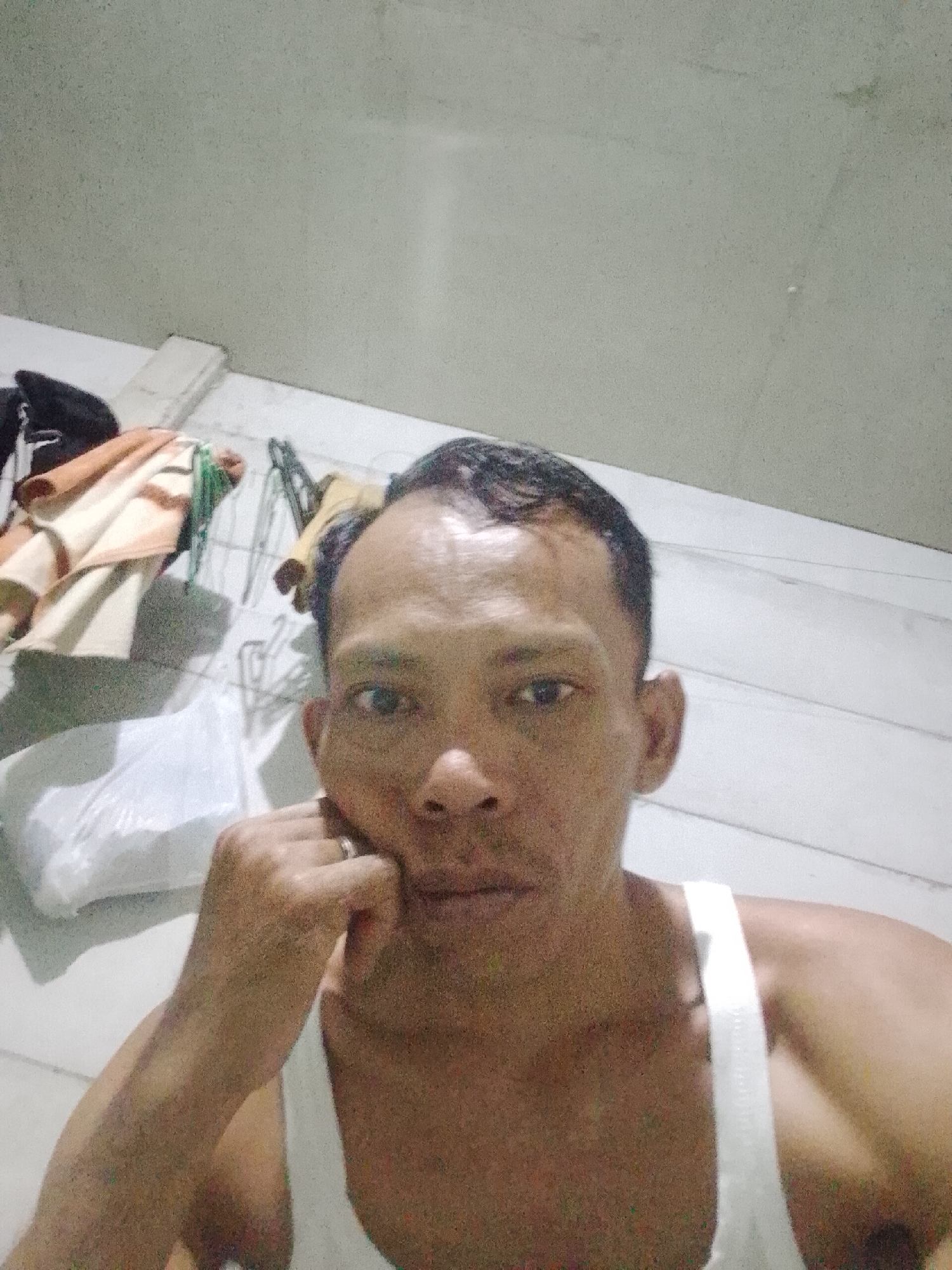 A man is in a room in Bukit Ketok, Indonesia. He is wearing a white tank top and resting his chin on his hand. There is a pile of clothes and a white plastic bag behind him on a tiled floor. The background is a plain light grey or white wall.