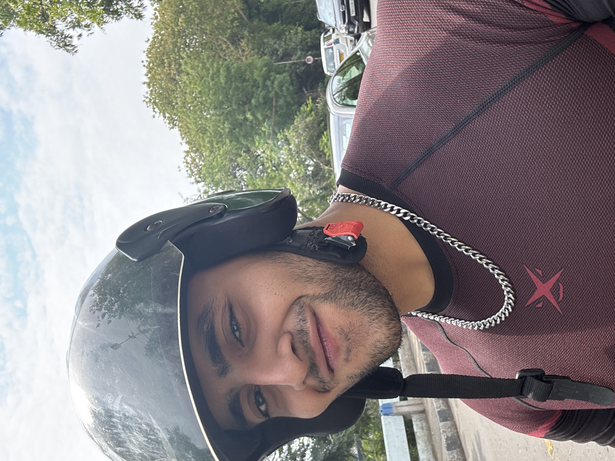 A person wearing a dark-colored helmet and a maroon-colored textured shirt with a silver chain around their neck is the focus of the image. The person has facial hair. The background shows a street scene, possibly in the city of Kolkata, India, with trees, parked cars, and infrastructure visible. The image is taken from a close range, focusing on the individual and the immediate surroundings.