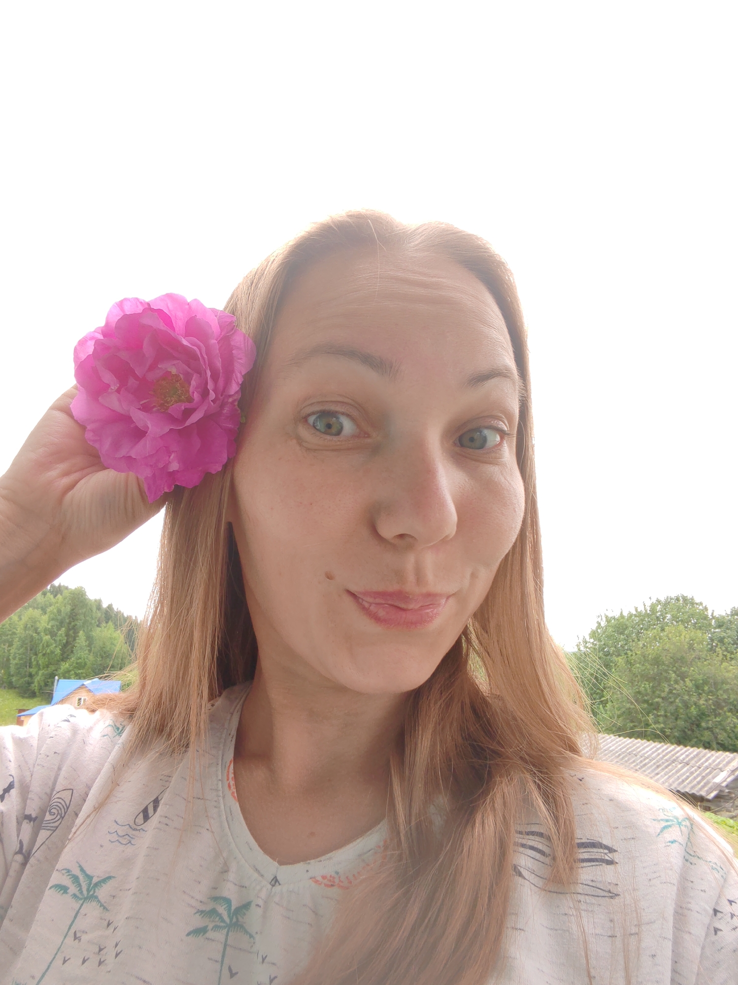 A woman with blonde hair is holding a pink flower behind her right ear. She is wearing a light-colored t-shirt with a tropical print. The background shows a bright sky, green trees, and a portion of a building with a corrugated roof. The scene appears to be outdoors in a rural setting, possibly in the city of Chukhlem, Russia.