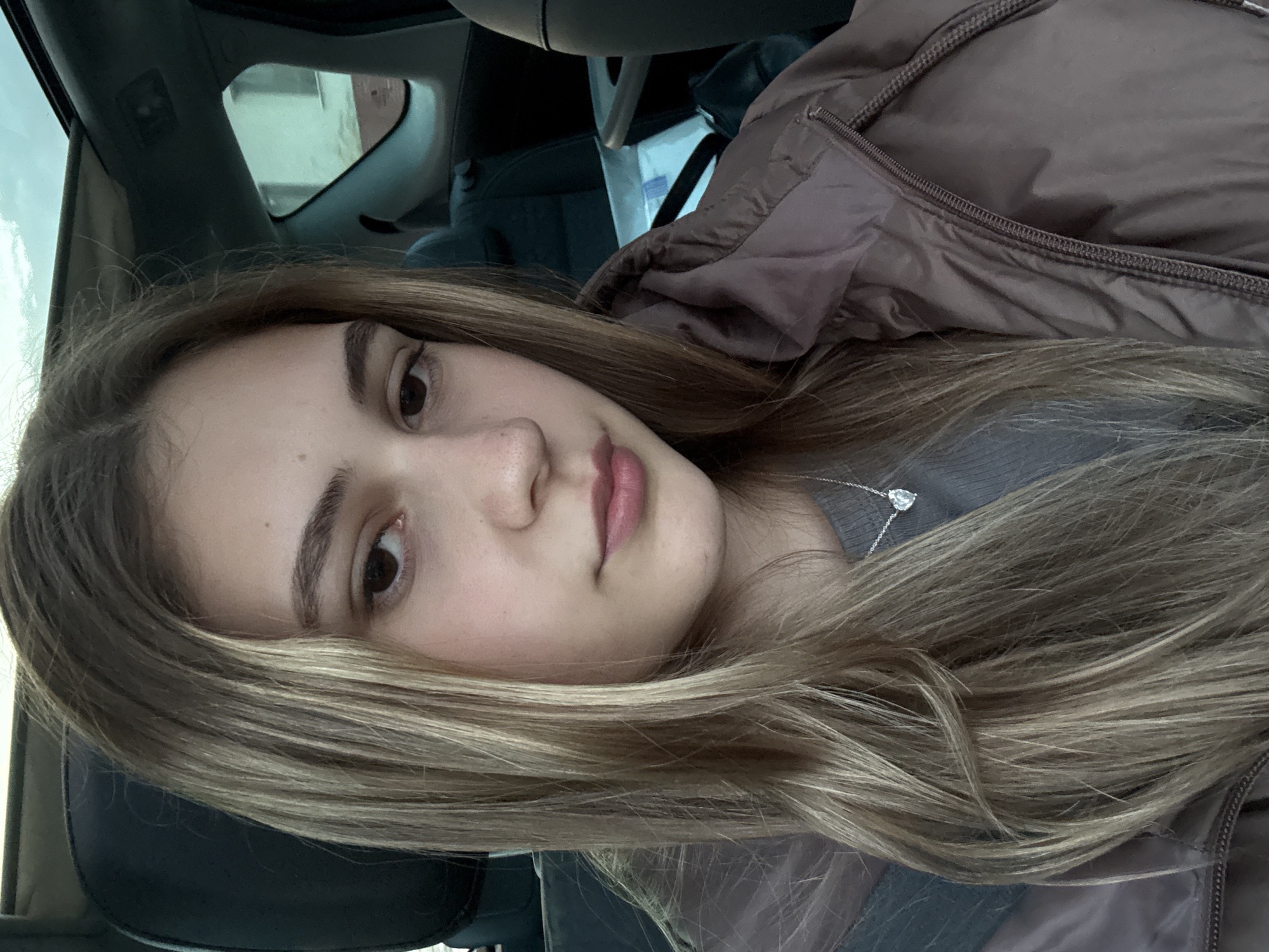 A medium close-up captures a young woman with light brown hair, fair skin, and brown eyes. She is wearing a gray top and a brown jacket, and a silver necklace with a heart pendant is visible. The woman is sitting inside a car, evident from the steering wheel and dashboard partially visible in the background. The lighting suggests daytime. The location and city of Panagyurishte, Bulgaria, cannot be ascertained from the given image.