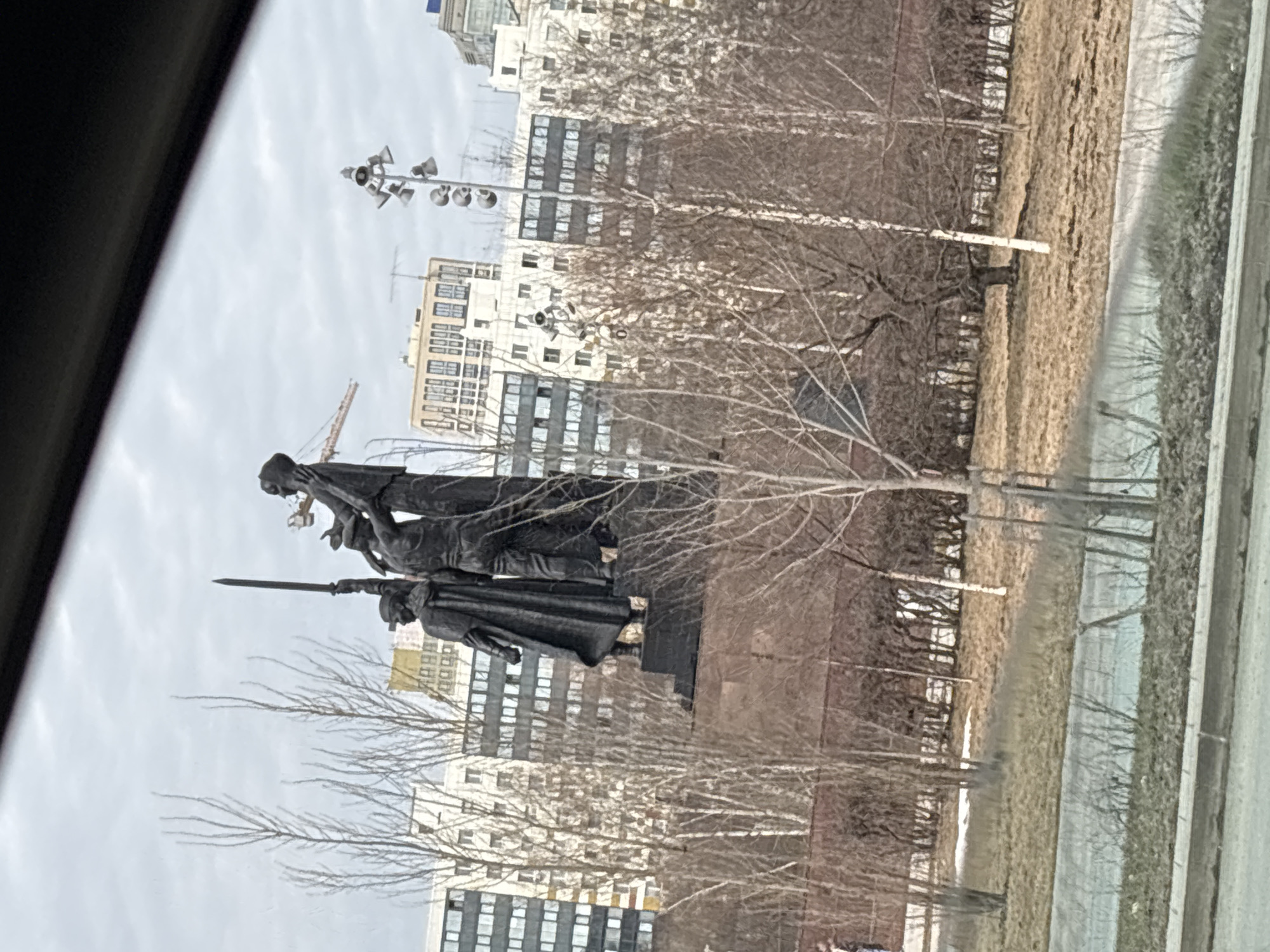 The image shows a view of a bronze monument through what appears to be a car window. The monument depicts three figures, one of whom holds a sword. The monument is placed in front of a backdrop of multi-story buildings and leafless trees, on a cloudy day. The location is believed to be the "Front to the Front" monument in Perm, Russia, next to the Kama River. A road or street is visible in the foreground.