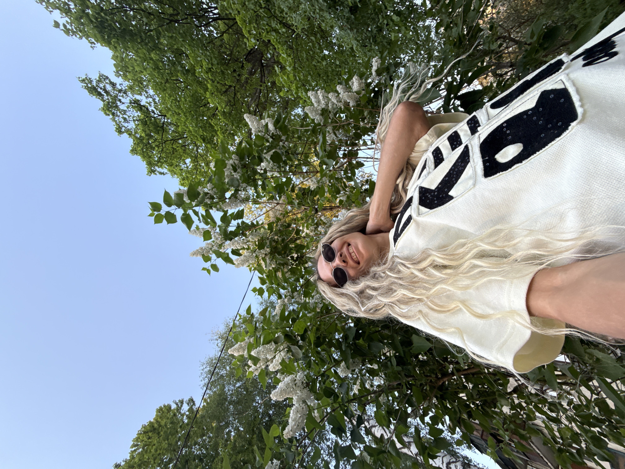 A woman with long blond hair and sunglasses is smiling at the camera. She is wearing a light-colored jacket with black patches. Behind her are trees with green leaves and white flowers, with a clear blue sky visible in the upper left. The photo appears to be taken from a low angle, looking up at the subject. Based on the visual information, the location in Rusanovo, Russia cannot be determined.