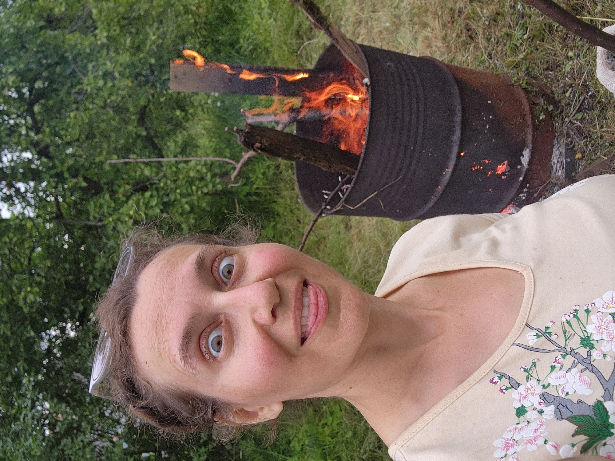 A medium shot photograph shows a woman with wide, surprised eyes and her tongue sticking out, looking directly at the camera. She has glasses perched on her head and is wearing a light-colored shirt with a floral design. Behind her, to the right, is a large, black metal barrel with a fire burning inside it, fueled by logs and branches. The barrel is situated on a grassy area with trees and greenery in the background. The scene appears to be outdoors in a rural or suburban setting.