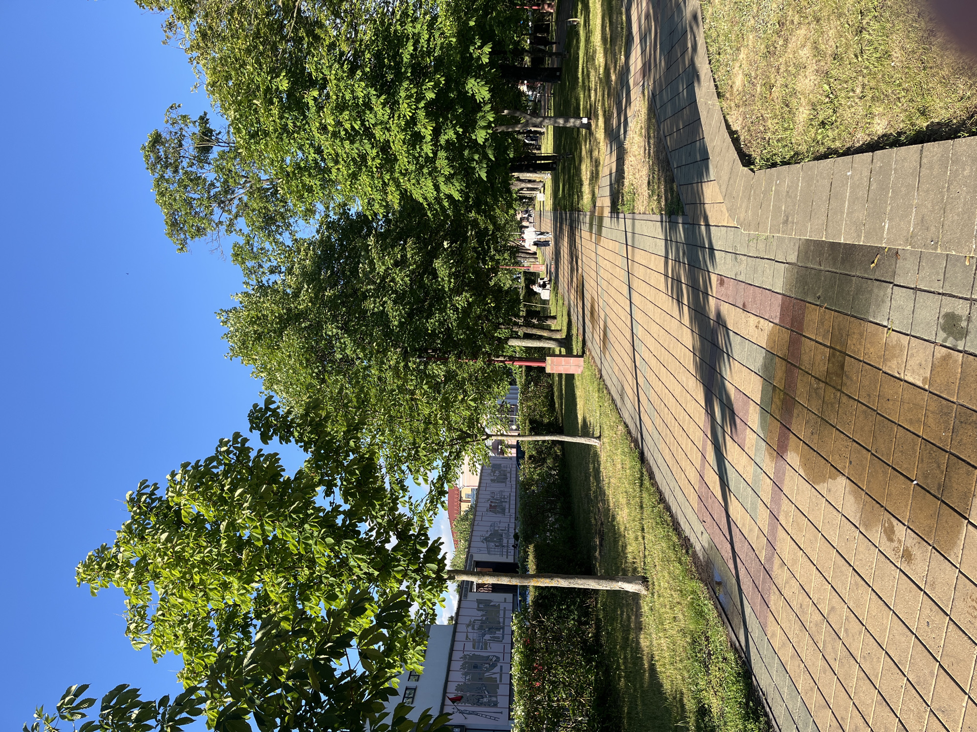 A park pathway with a patterned paved surface and a grass area on the left is shown under a clear blue sky on a sunny day. The pathway is lined with trees that cast shadows on the ground. In the background, there is a building with a mural and a few people walking along the path.