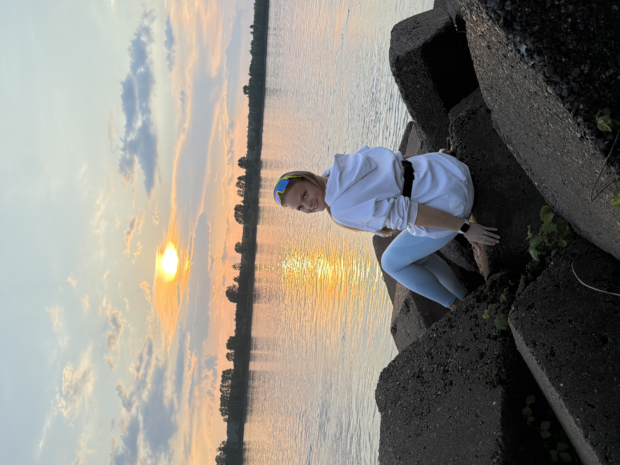 A young woman with blonde hair is crouching on large, dark, rough-textured concrete blocks by the water. She is wearing a white hoodie, light blue leggings, and brightly colored sports glasses pushed up on her forehead. A black belt is cinched around her waist. Her right hand is resting on the concrete blocks, and her left hand is near her leg, with a smartwatch visible on her wrist. She is smiling and looking towards the camera.
The scene is a body of water, likely a lake or river, with gentle ripples reflecting the sky. On the far bank, a line of trees is silhouetted against the sky. The sun is low in the sky, casting a warm, golden glow on the water and illuminating the clouds, suggesting it is either sunrise or sunset. The sky is a mix of pale blue and soft oranges and yellows, with scattered clouds. The overall mood is peaceful and serene, with a touch of warmth from the golden hour light.
The location is identified as Chaikovsky, Russia. No visible text is present in the image.