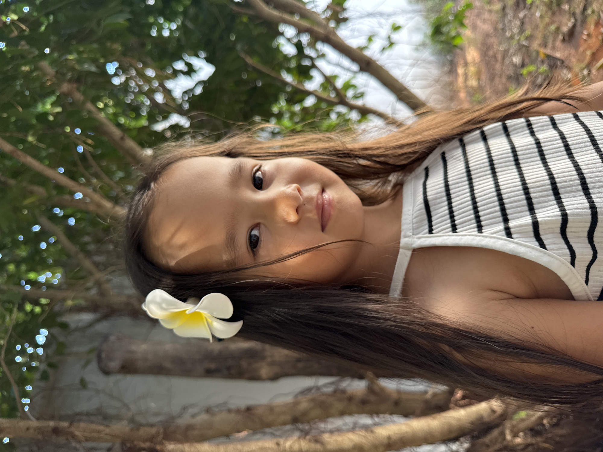 A young girl with long brown hair is the primary subject of this portrait-style photograph taken in Mũi Né, Vietnam. A white and yellow plumeria flower is positioned in her hair. She wears a white tank top with black vertical stripes. The backdrop includes tree branches and green foliage, along with blurred white lights suggesting a possibly bright or sunlit environment. The overall perspective is angled, with the ground and tree branches appearing in the foreground.